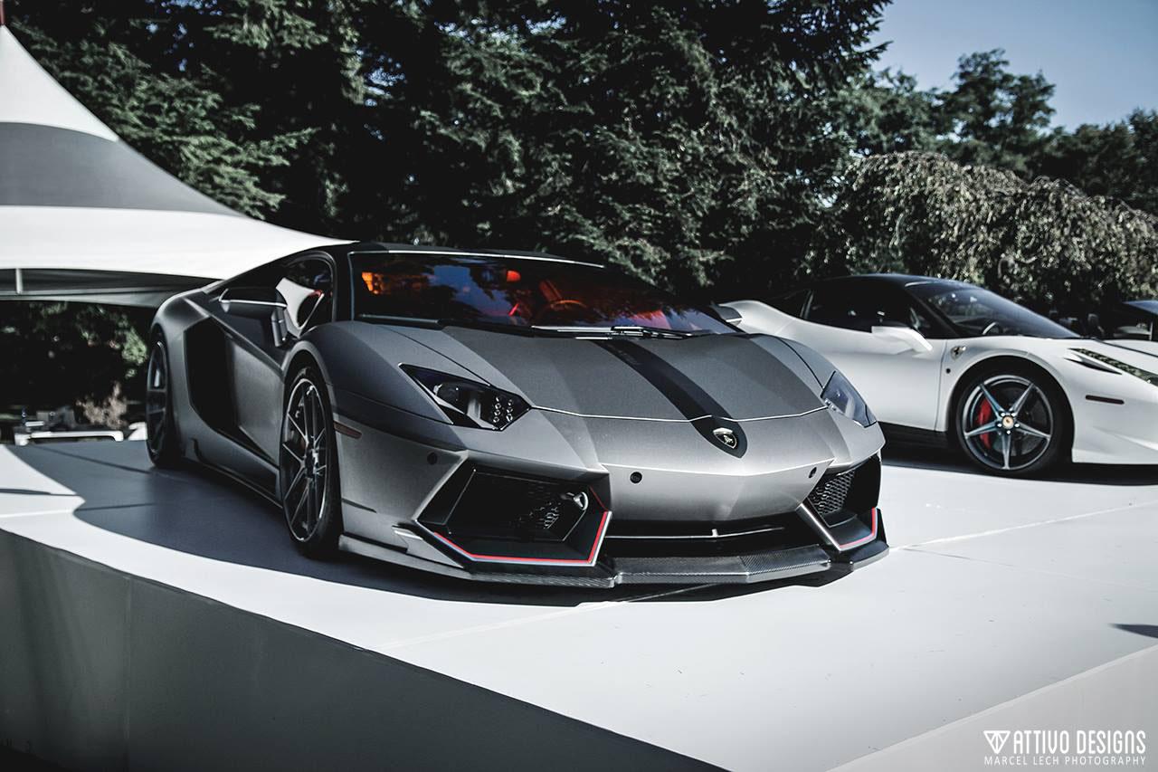 Front three-quarter view of a matte grey Lamborghini Aventador with a black racing stripe and red accents, displayed on a white platform next to a white Ferrari.