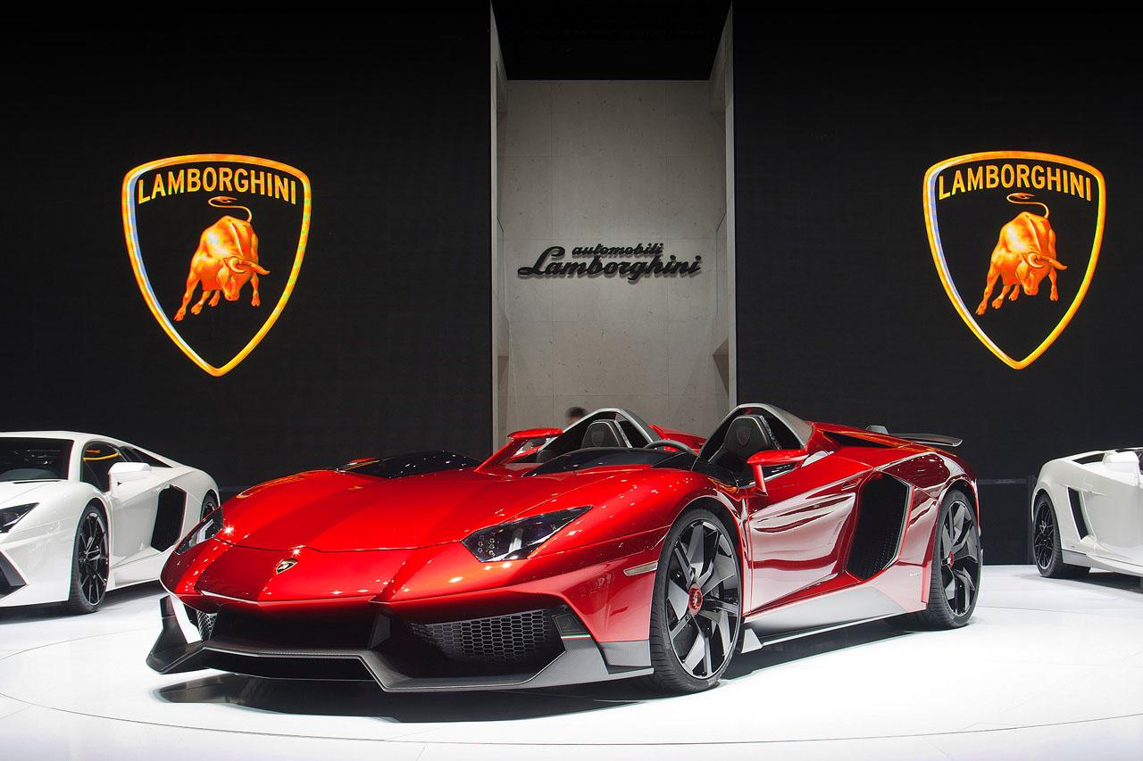 The striking red Aventador J displayed prominently at an auto show, flanked by two white Aventadors, against a dramatic Lamborghini branded backdrop.