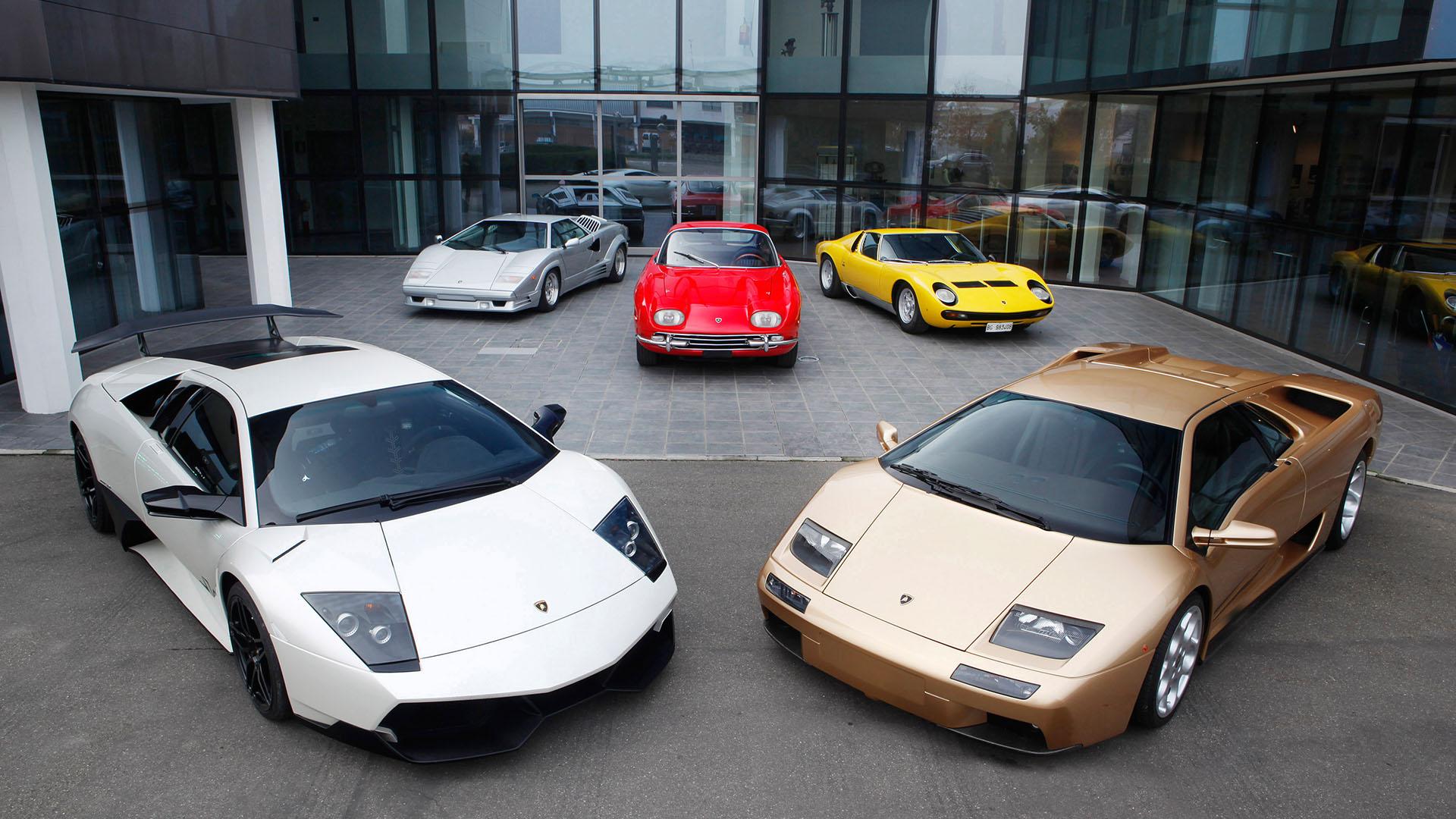 A collection of six significant Lamborghini models from different eras displayed in front of a modern glass building, likely the factory or museum.