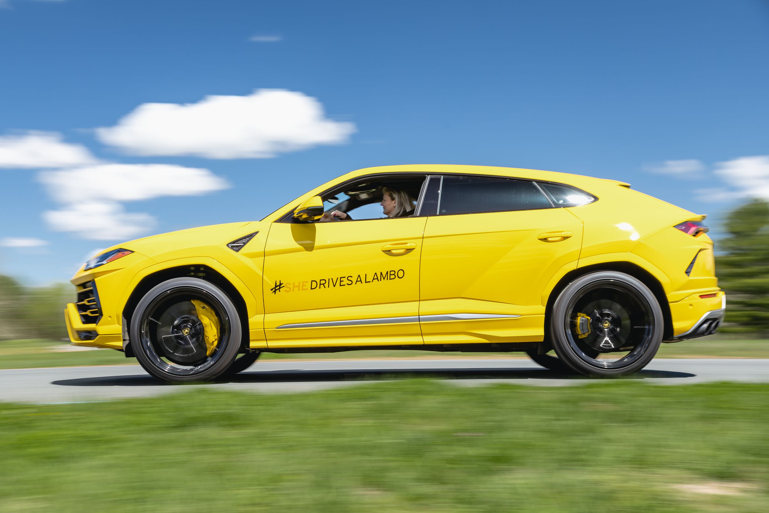 A bright yellow Lamborghini Urus is captured in motion on a scenic road, with a female driver visible, featuring the '#SHEDRIVESALAMBO' decal.