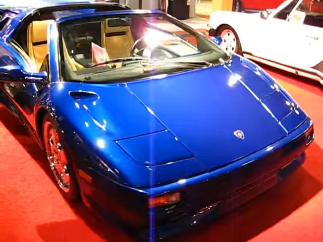A vibrant metallic blue Lamborghini Diablo VT Roadster displayed on a red carpet at an indoor car show or exhibition.
