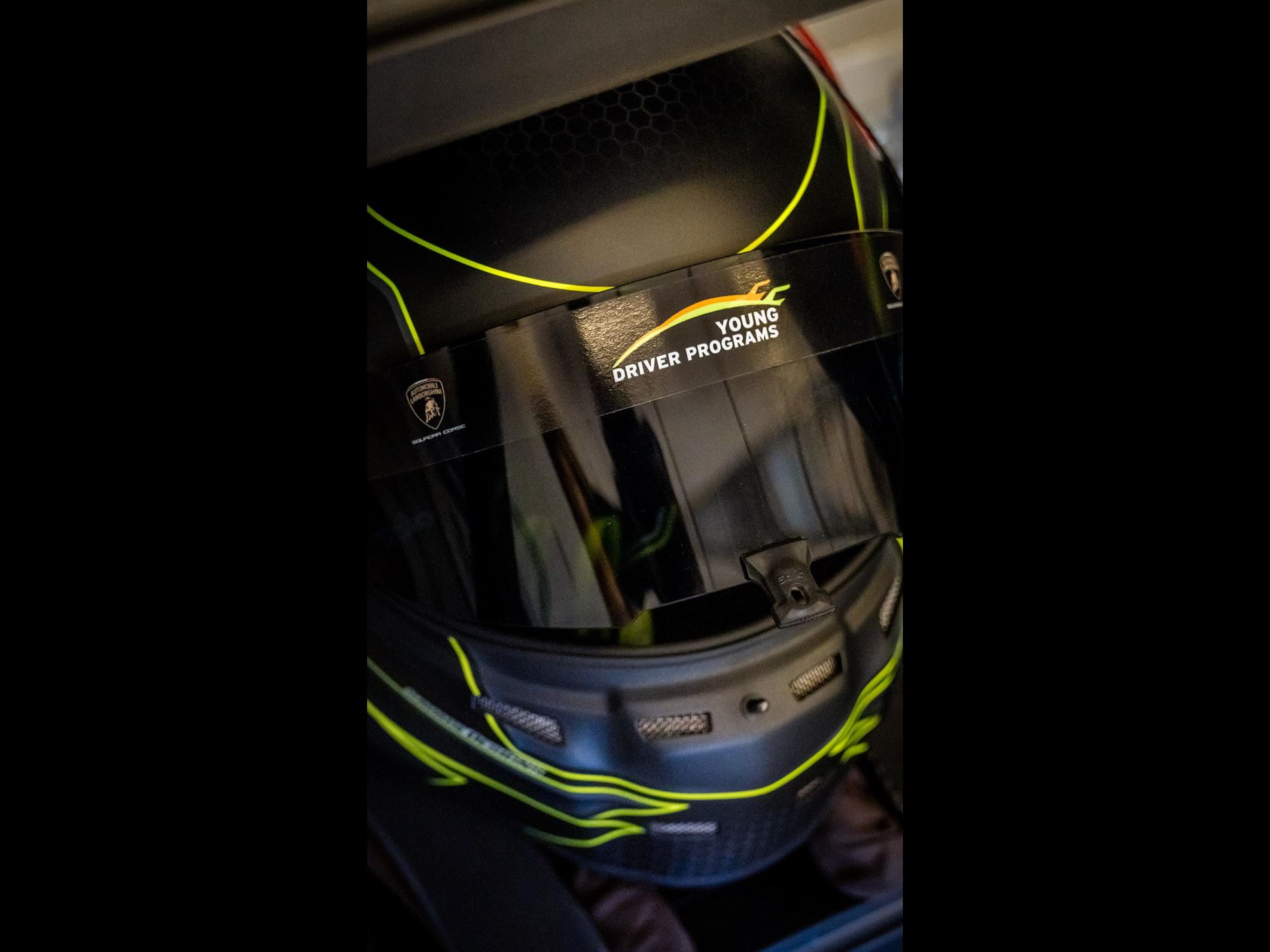 Lamborghini'S Talent Pipeline Delivers: Paul And Michelotto Earn Factory Seats, Cook And Queen Named Junior Winners 15 A detailed shot of a black racing helmet with bright yellow accents and the 'young driver programs' logo. The helmet's design suggests a focus on safety and performance, crucial for aspiring racing ta