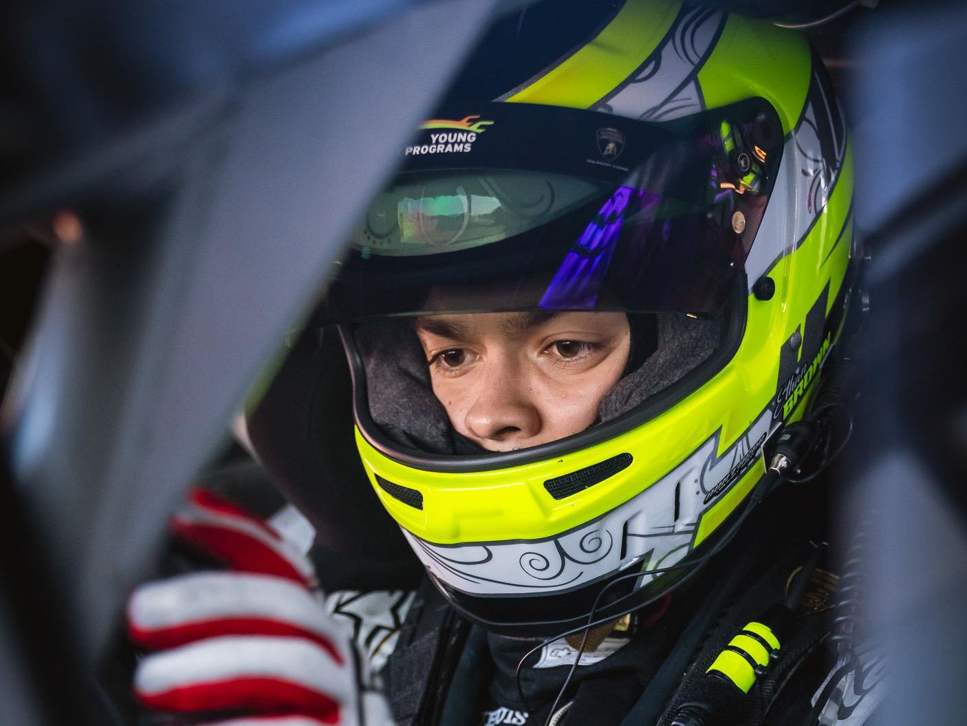 Lamborghini'S Talent Pipeline Delivers: Paul And Michelotto Earn Factory Seats, Cook And Queen Named Junior Winners 17 A close-up of a young racing driver in a helmet, peering intently from the cockpit of a race car. The helmet's bright yellow and white design, along with the 'young driver programs' logo, signifies th