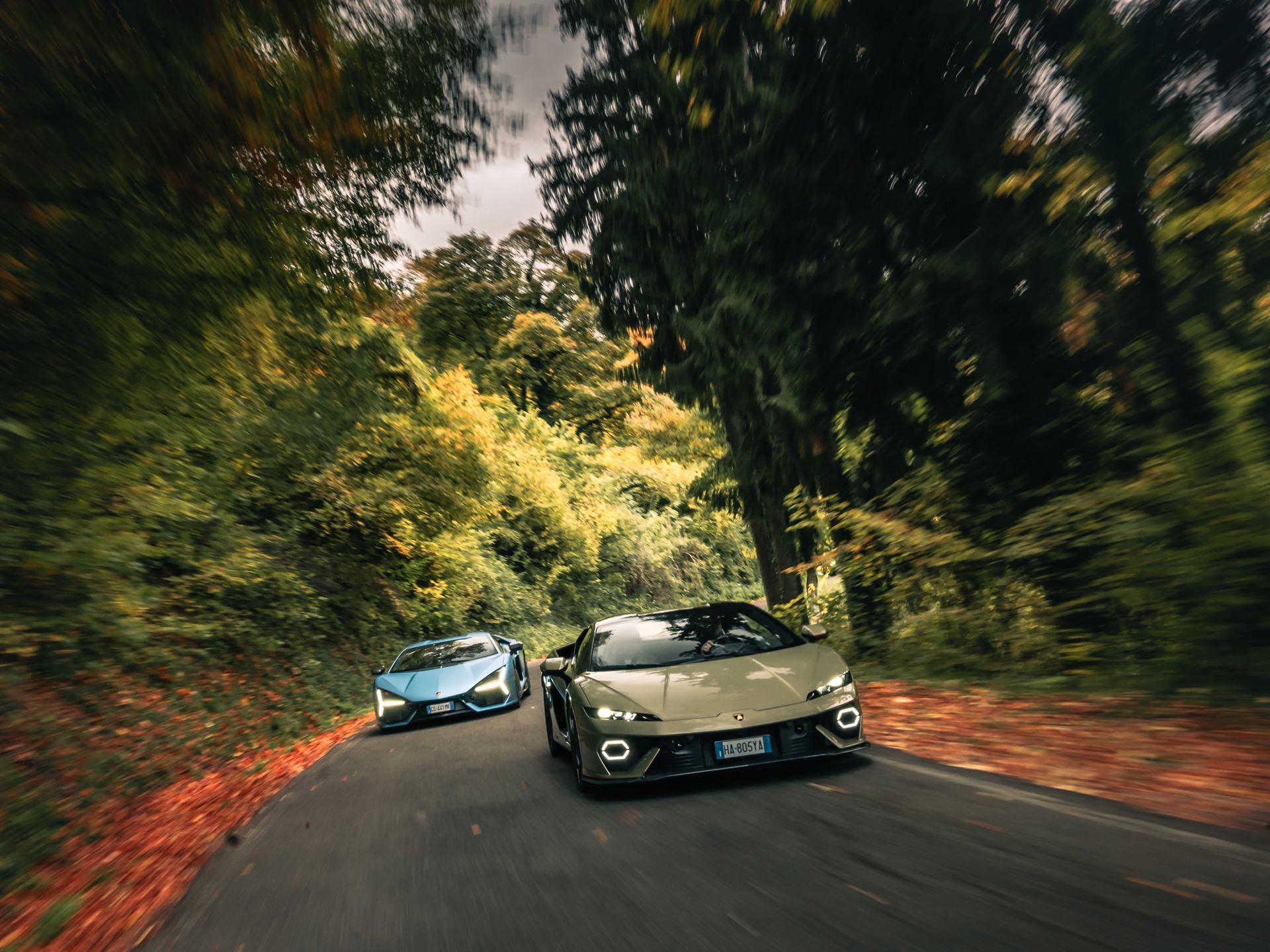Two lamborghini huracán models (one beige/gold, one light blue) driving dynamically on a winding road with autumn foliage, creating a sense of speed with motion blur.