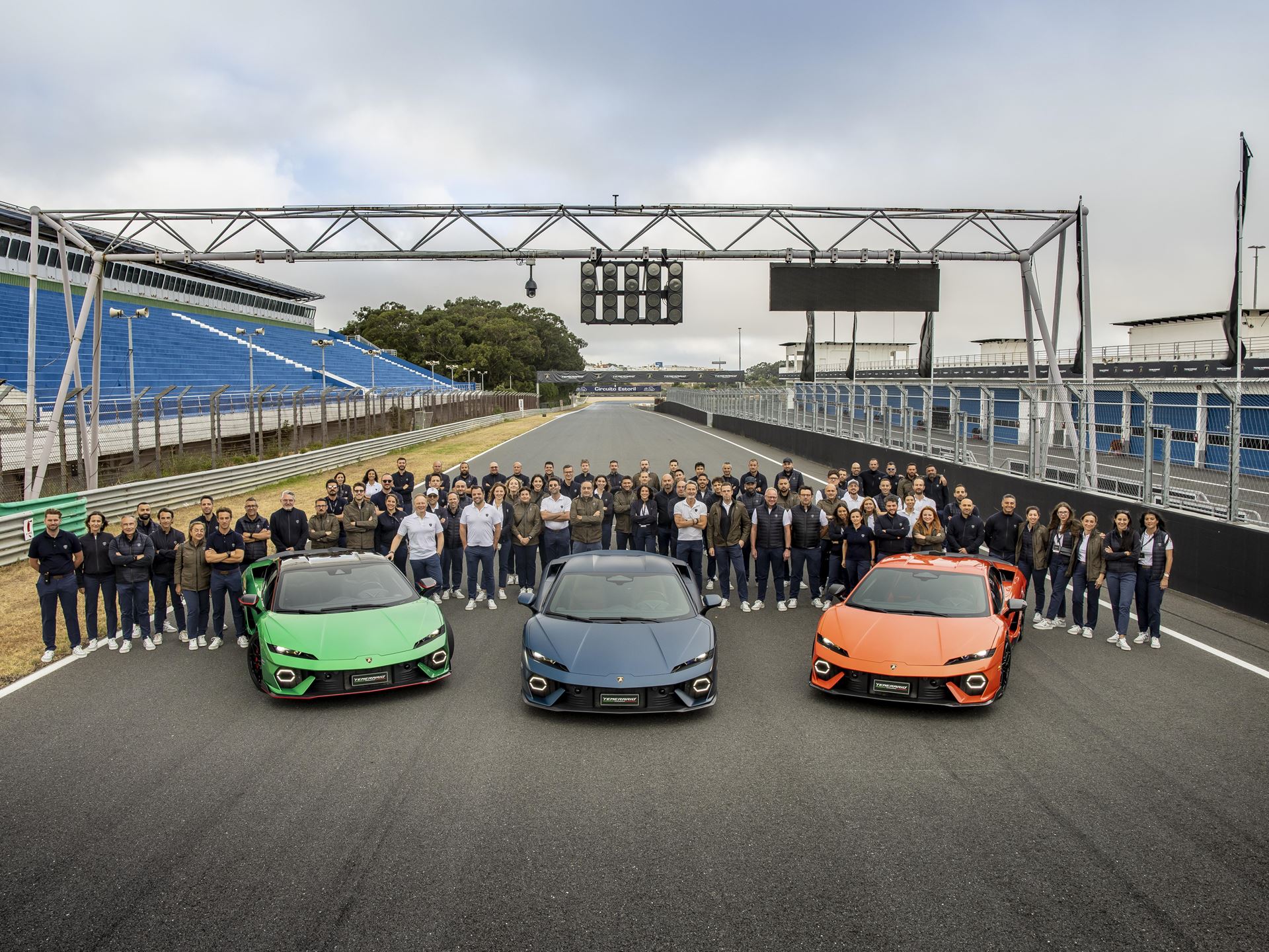 Three lamborghini huracán sterrato models (green, blue, orange) lined up on a racetrack, with a large group of people behind them and a starting lights structure overhead.