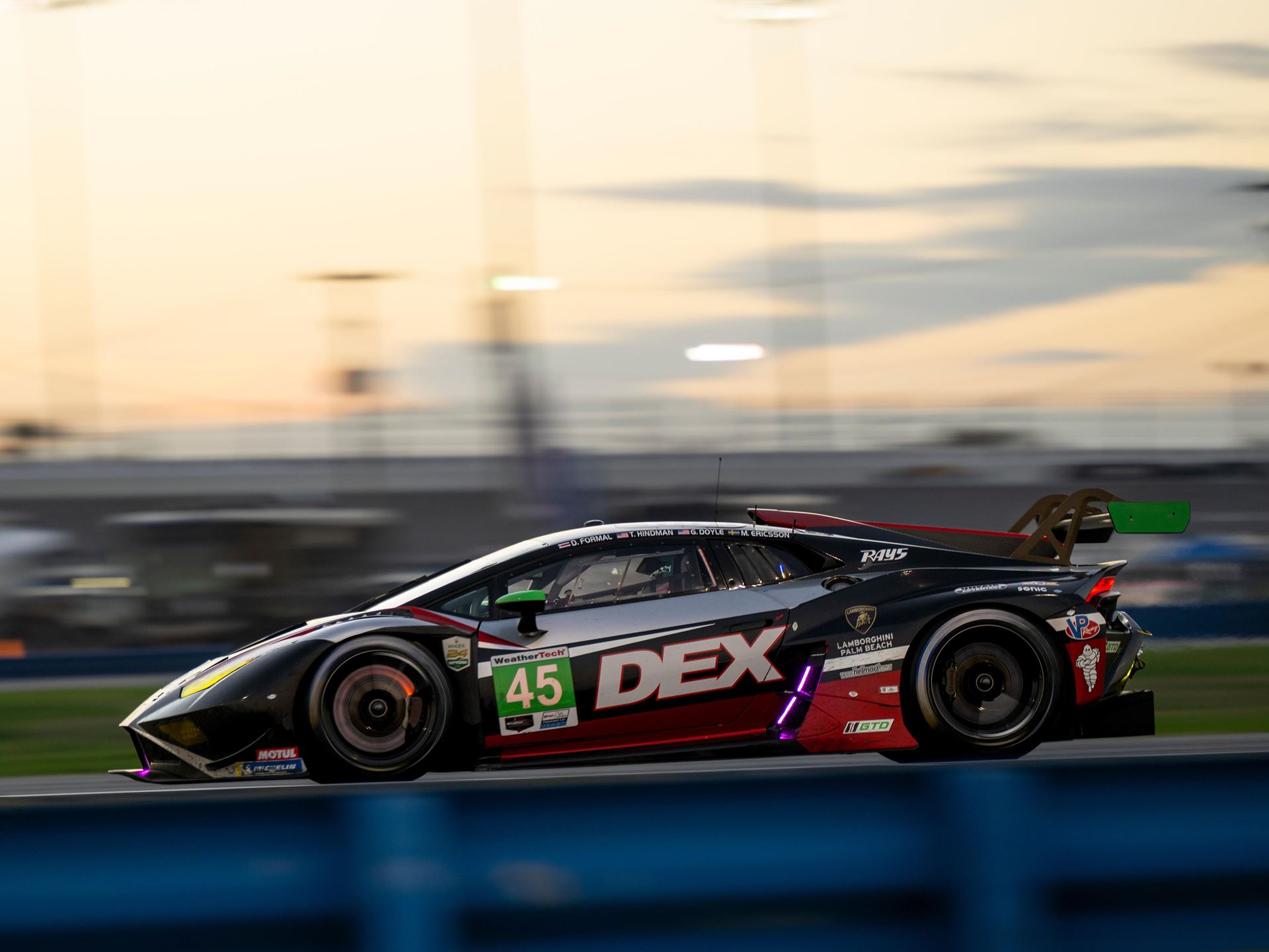 Lamborghini'S First In-House Gt3 Car Faces Brutal Sebring Debut 17 A lamborghini huracan gt3 evo race car, number 45, speeds along a race track during the golden hour, with motion blur emphasizing its velocity. The car's black, white, and red livery with green accent