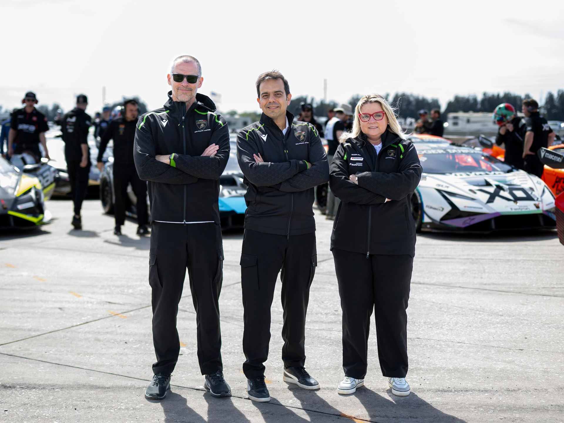 Lamborghini'S New Racing Boss Comes Straight From Ferrari'S Playbook, And The Timing Is No Coincidence 7 Three individuals, two men and one woman, wearing black lamborghini squadra corse jackets, stand with their arms crossed on a track. Behind them, several lamborghini huracan race cars are parked, with