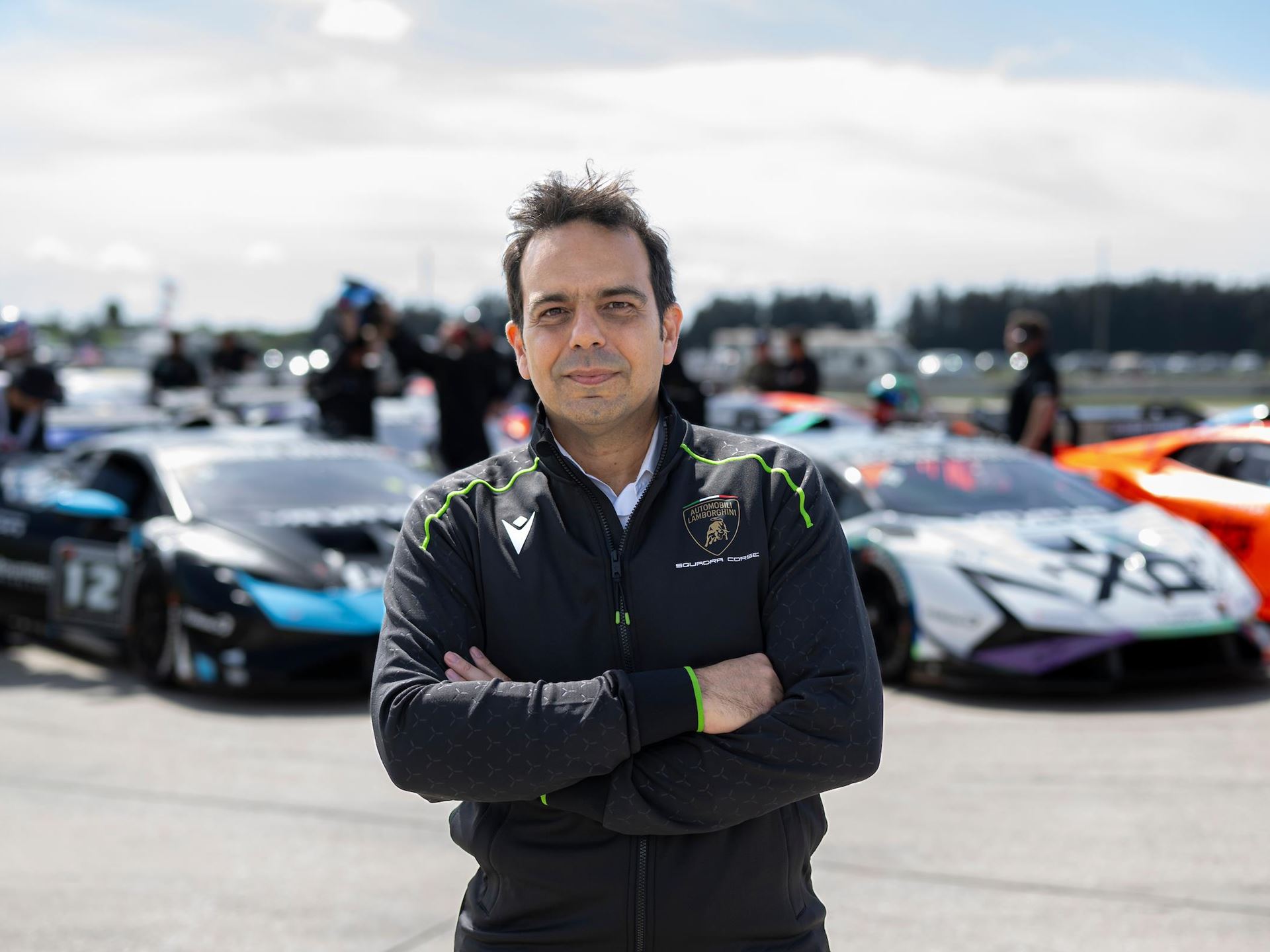 Lamborghini'S New Racing Boss Comes Straight From Ferrari'S Playbook, And The Timing Is No Coincidence 9 A man in a black lamborghini squadra corse jacket stands with his arms crossed, smiling slightly at the camera. In the background, several lamborghini huracan race cars are visible on a track under a