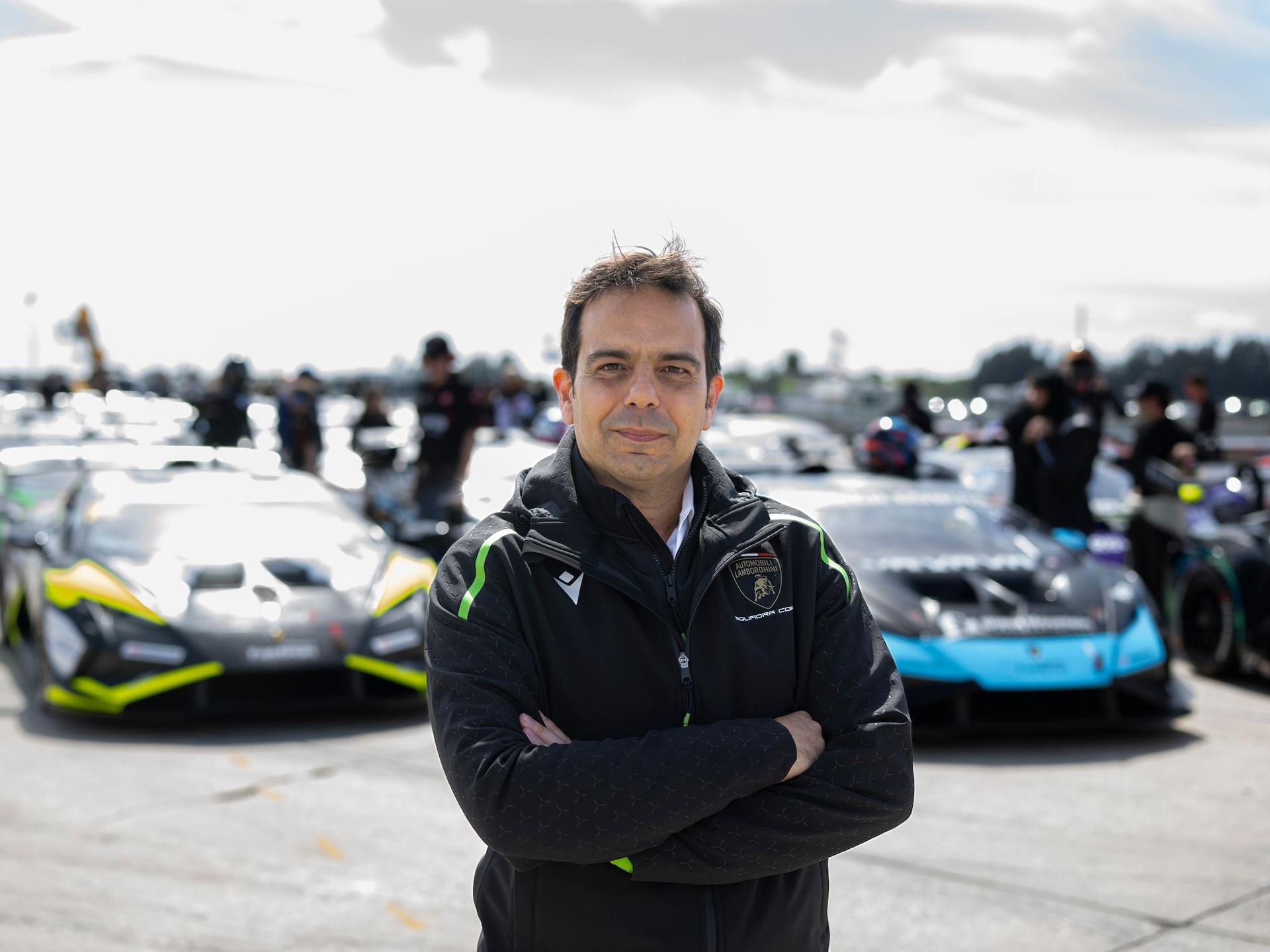 Lamborghini'S New Racing Boss Comes Straight From Ferrari'S Playbook, And The Timing Is No Coincidence 11 A man in a black lamborghini squadra corse jacket stands with his arms crossed, looking directly at the camera. Behind him, a line of lamborghini huracan race cars are parked on a track, suggesting a