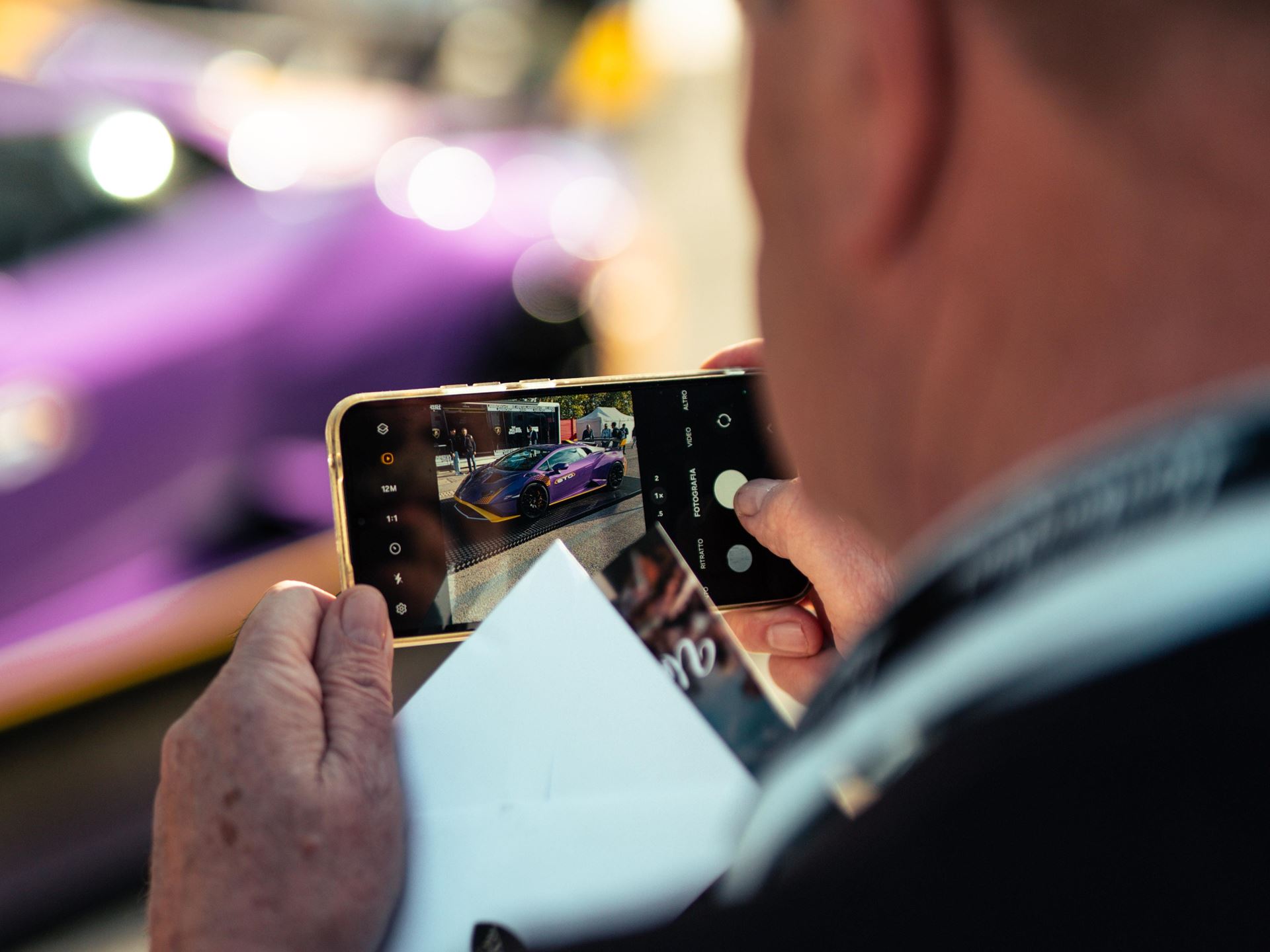 Lamborghini Arena 2026 Brings Live Super Trofeo Racing To Imola, Plus The Temerario Race Car'S Public Debut 9 A person is seen from behind, holding a smartphone and taking a picture of a vibrant purple lamborghini huracan sto. The car is clearly visible on the phone screen, showcasing its distinctive design a