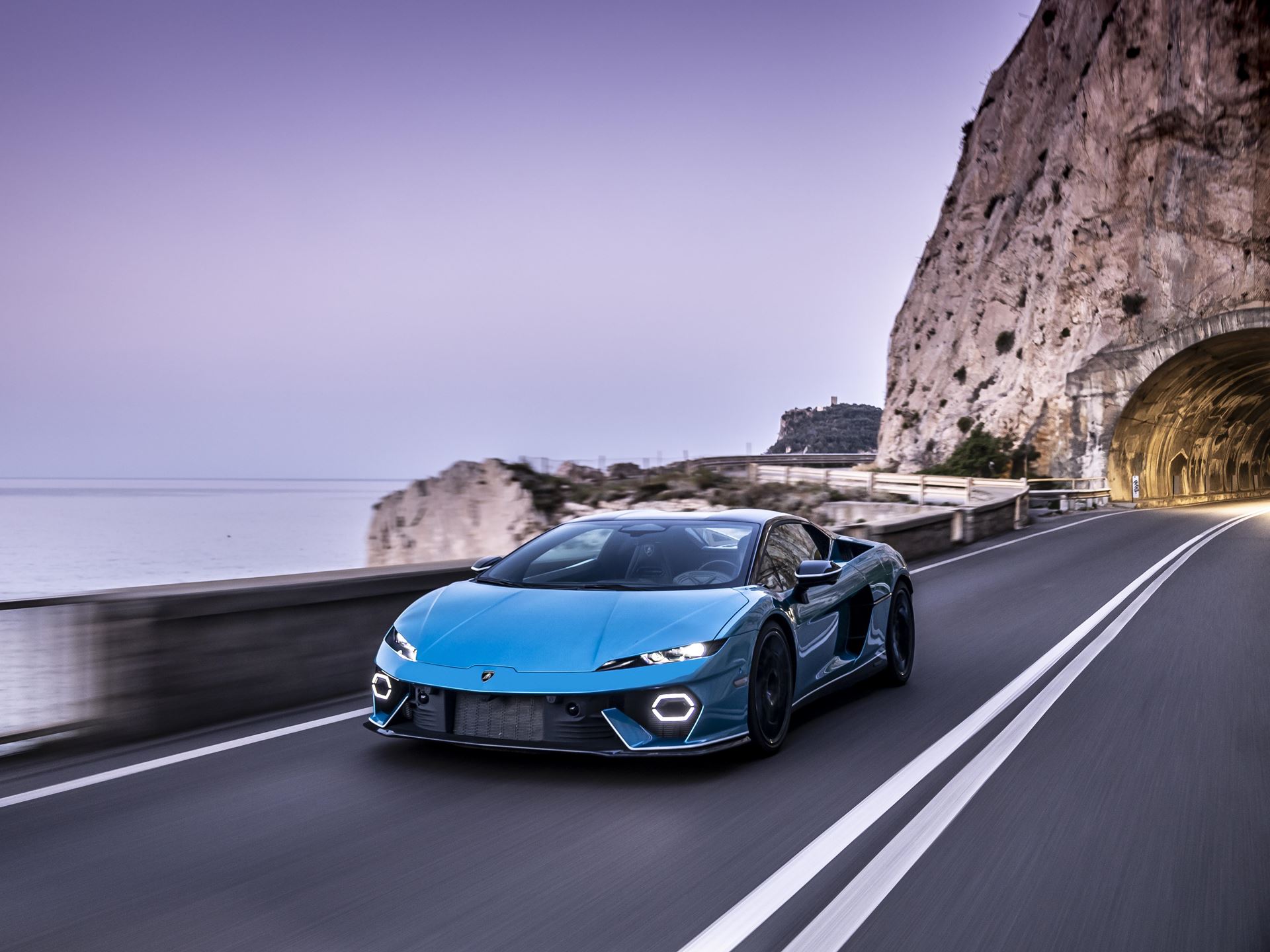 A blue lamborghini temerario driving along a scenic coastal road with a tunnel and ocean views.