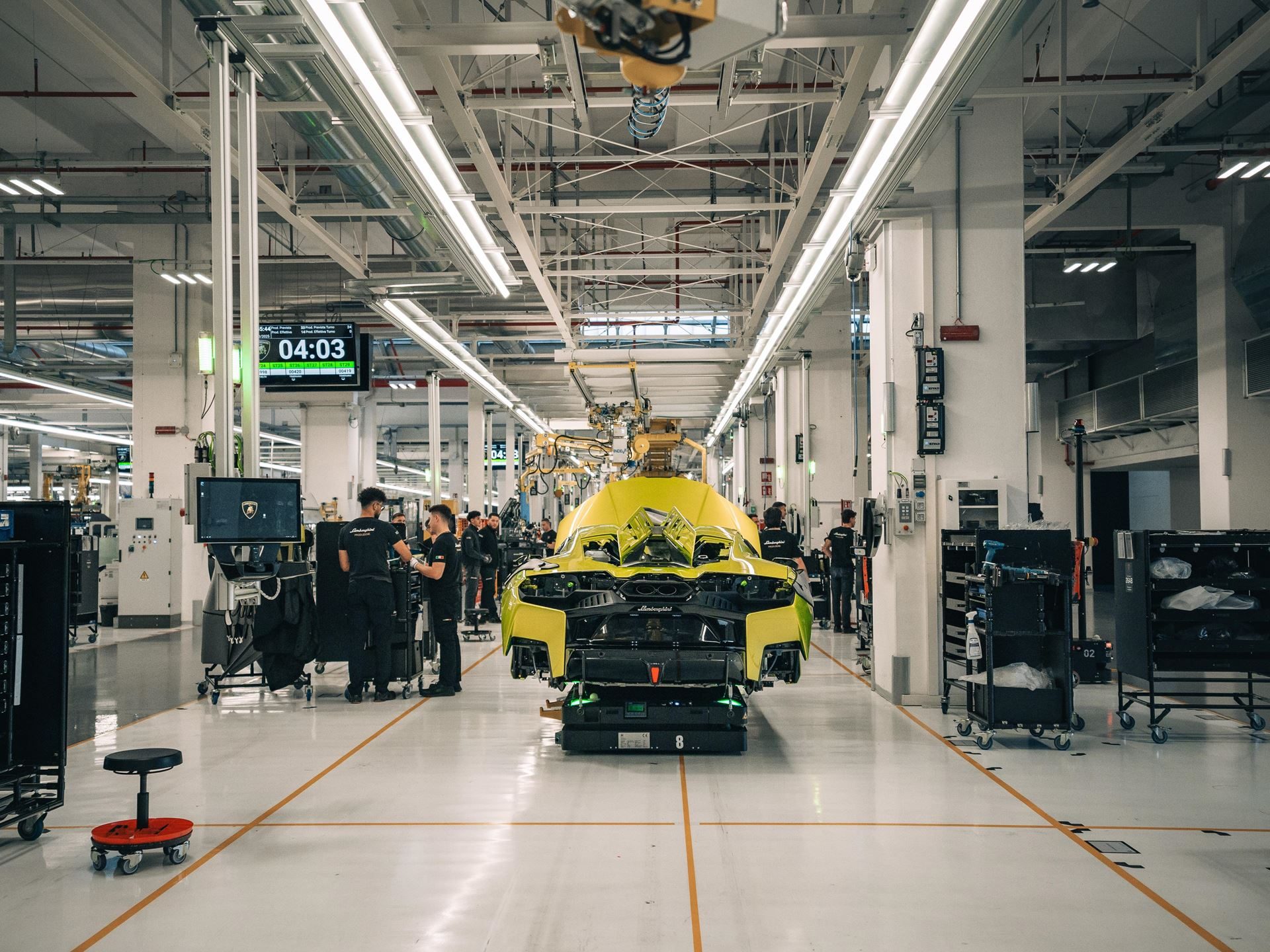 A yellow Lamborghini chassis on the assembly line with technicians working around it in a bright factory environment
