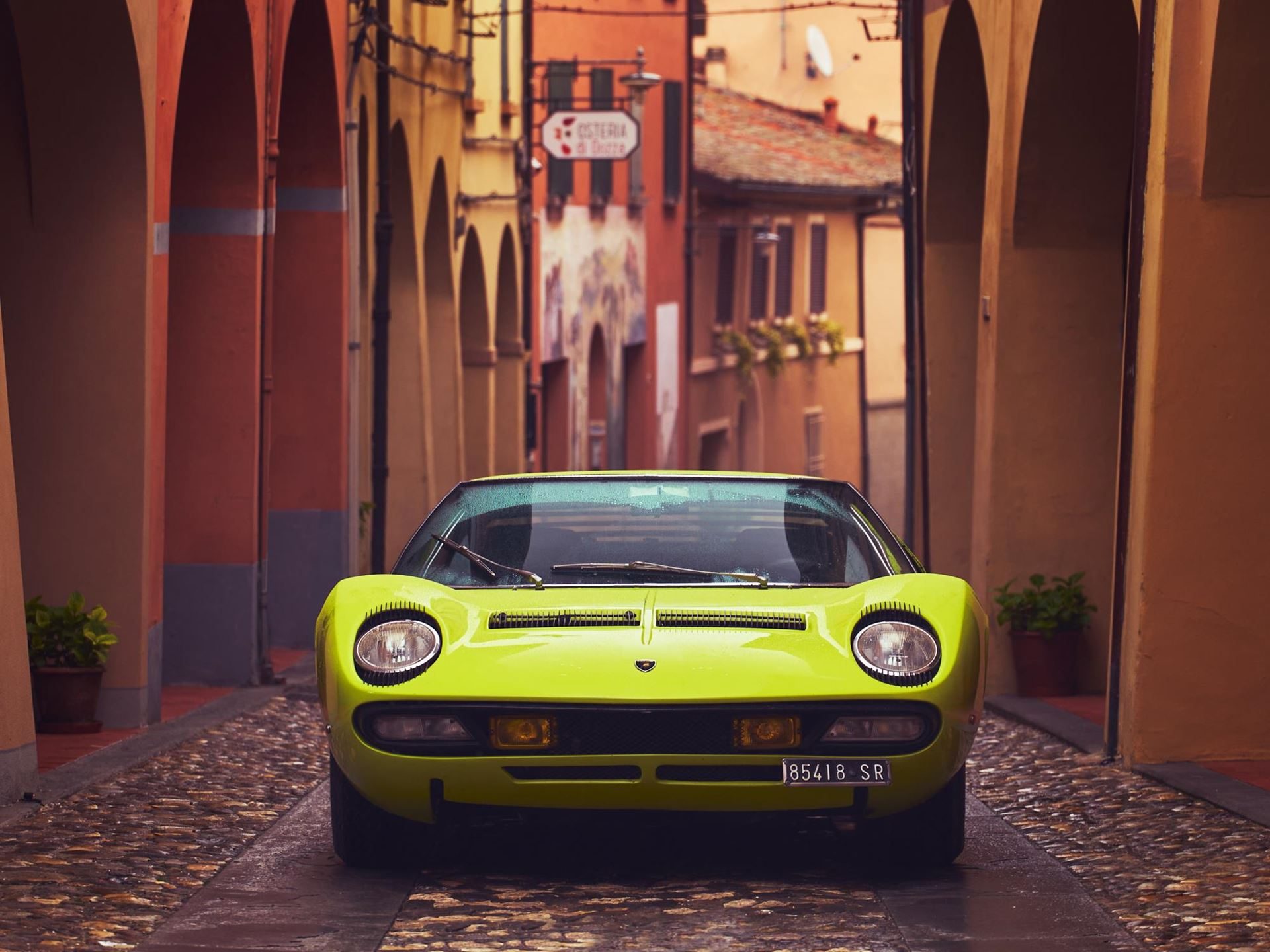 The Counterclockwise Crankshaft And Shared Sump That Invented The Supercar: 60 Years Of Lamborghini Miura 5 Vibrant yellow lamborghini miura parked on a cobblestone street framed by historic italian archways, showing its front view, pop-up headlights, and low-slung profile