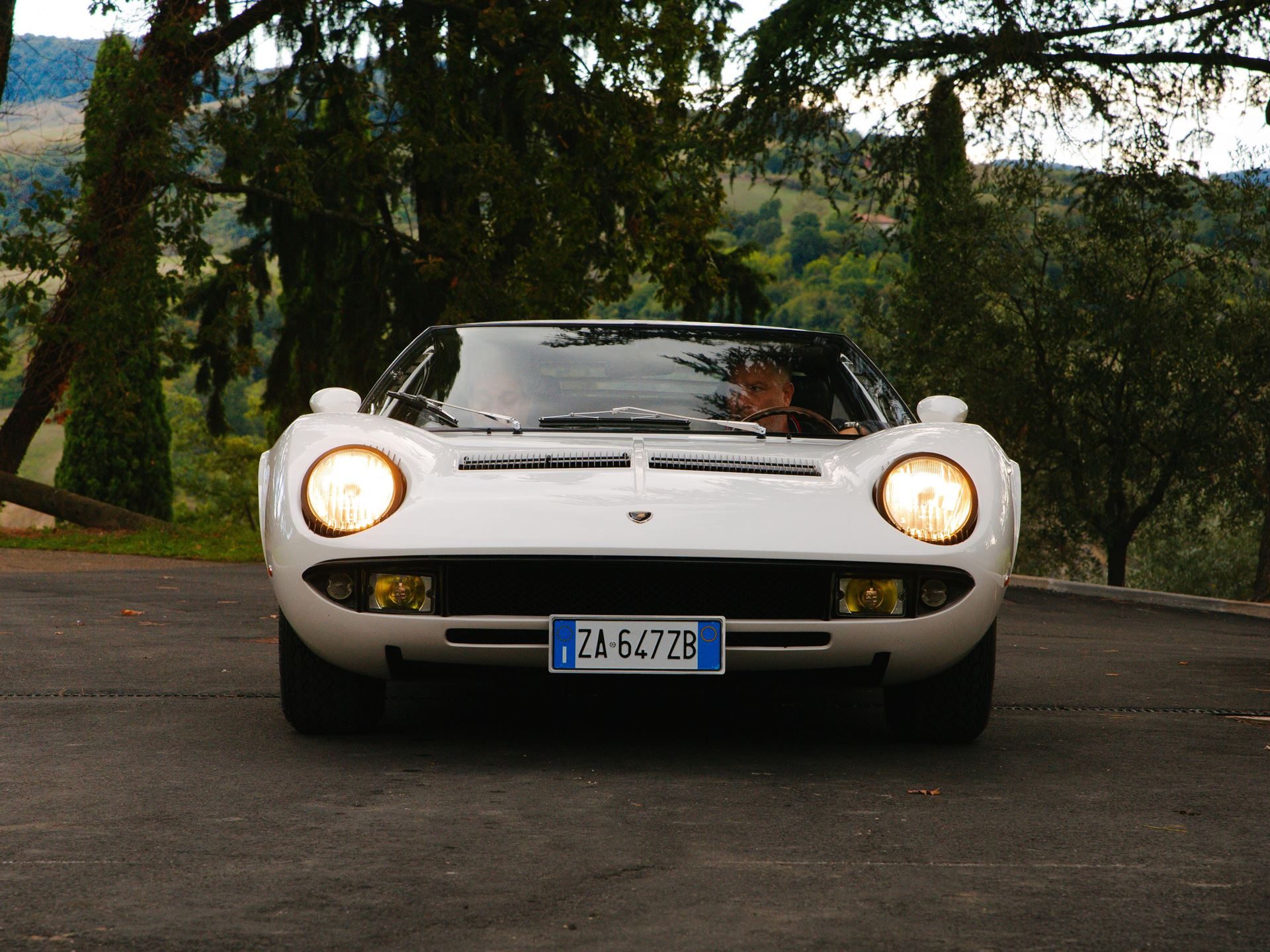 The Counterclockwise Crankshaft And Shared Sump That Invented The Supercar: 60 Years Of Lamborghini Miura 9 White lamborghini miura in a head-on shot with pop-up headlights illuminated, driver partially visible, against a natural backdrop of trees