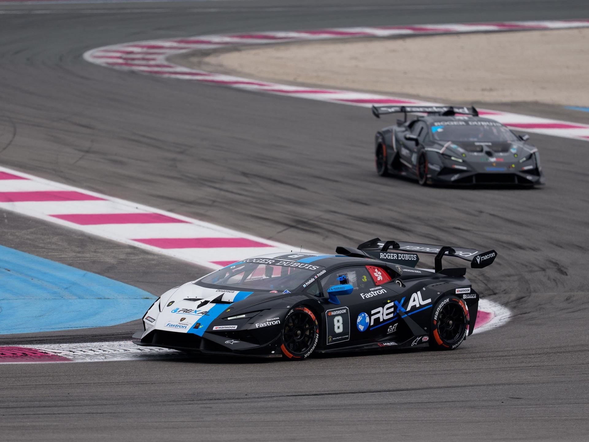 A black lamborghini huracán super trofeo evo2 with blue and white accents leads another car through a paul ricard turn, both showing aggressive bodywork and large rear wings