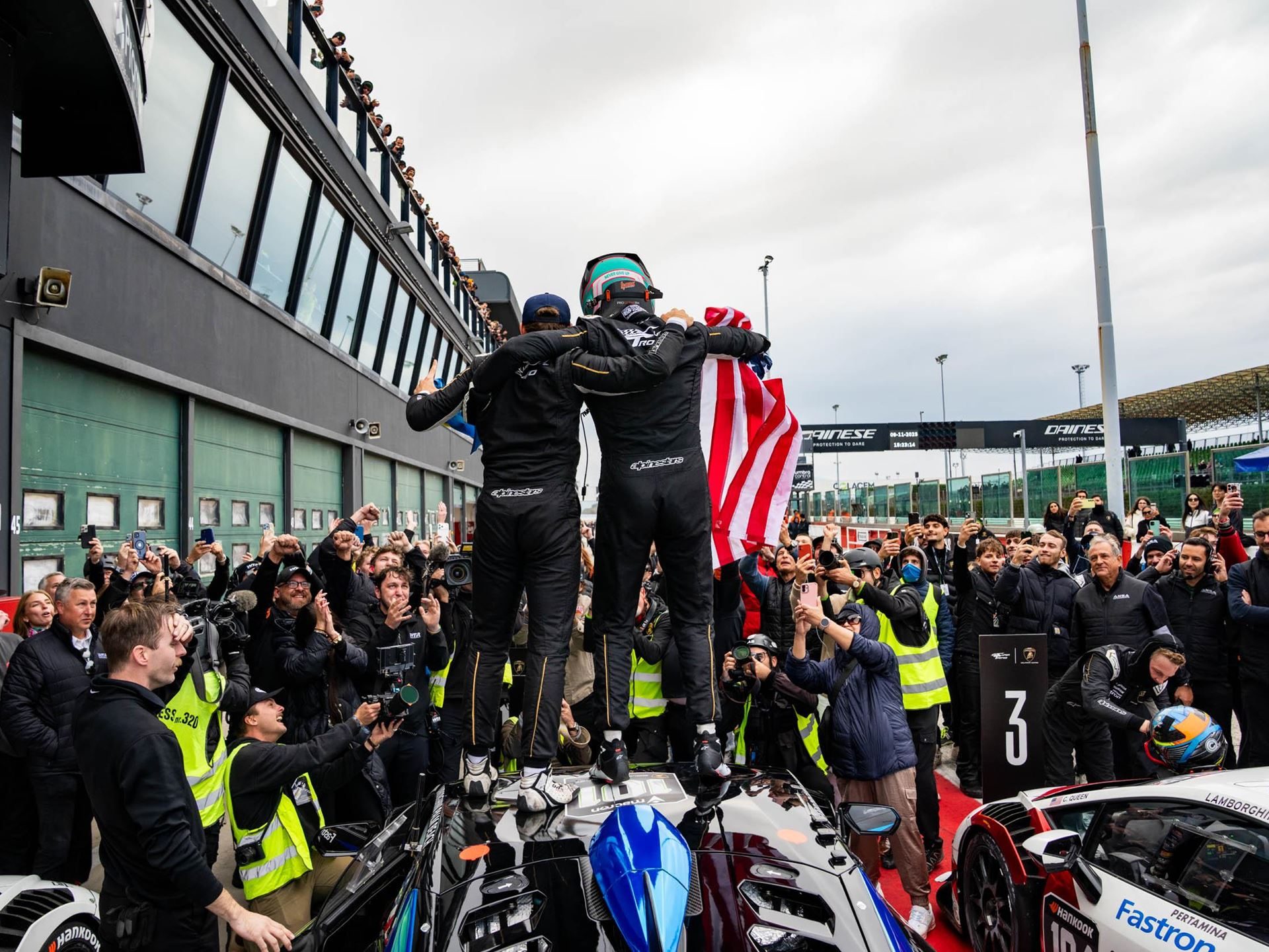 Eleven Winners, Seven Champions: The Huracán Super Trofeo Evo2'S Triumphant Farewell At Misano 5 Two race car drivers celebrating atop a lamborghini huracán race car, holding an american flag amid a cheering crowd at the world finals