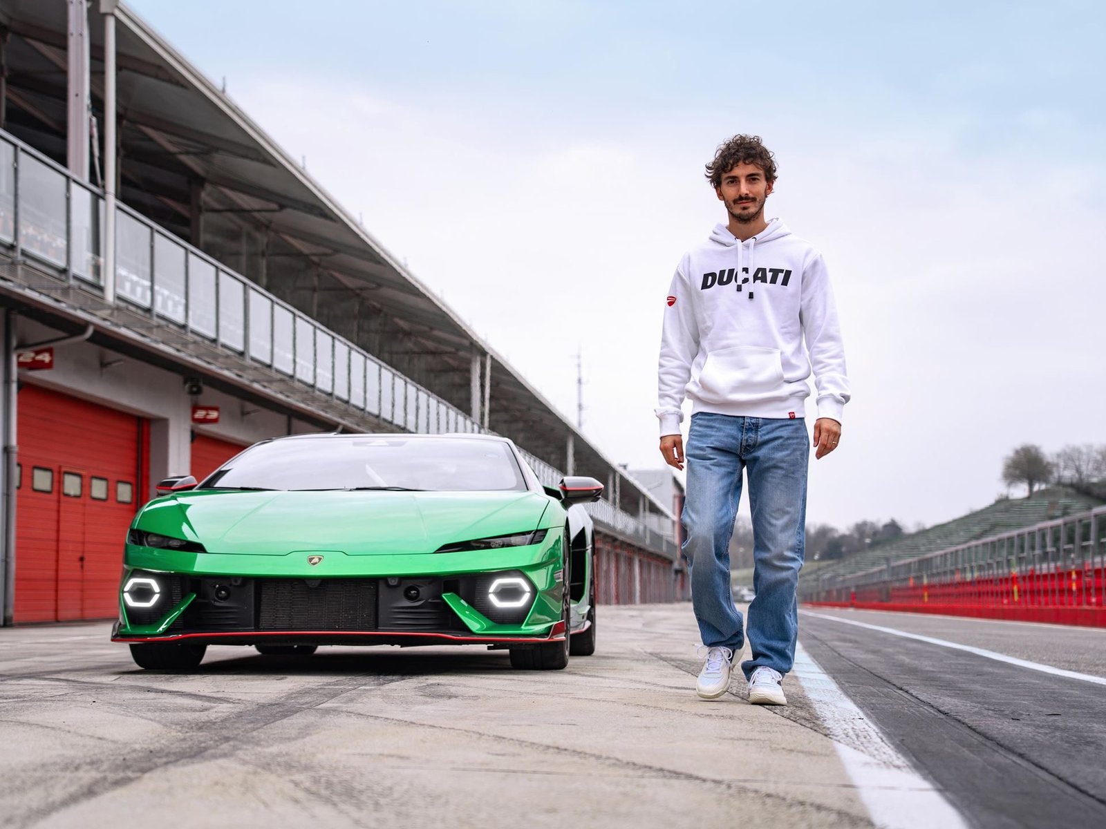 Francesco Bagnaia walks toward the camera with a green Lamborghini Temerario positioned behind him on a racetrack pit lane, its Y-shaped DRLs and wide stance clearly visible