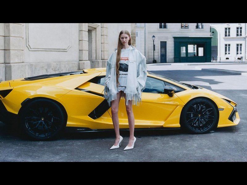 Female model in Balenciaga x Lamborghini apparel standing beside a yellow Lamborghini Revuelto on a city street