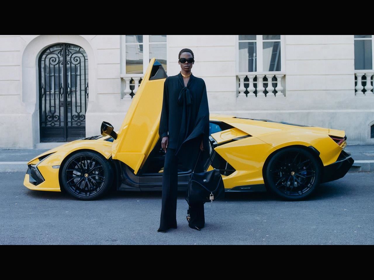 Balenciaga Lamborghini Collaboration Puts Temerario Artwork On The Runway And Graffiti-Wrapped Revueltos Outside Flagships Worldwide 3 Female model in black balenciaga x lamborghini attire in front of a yellow lamborghini revuelto with scissor door open