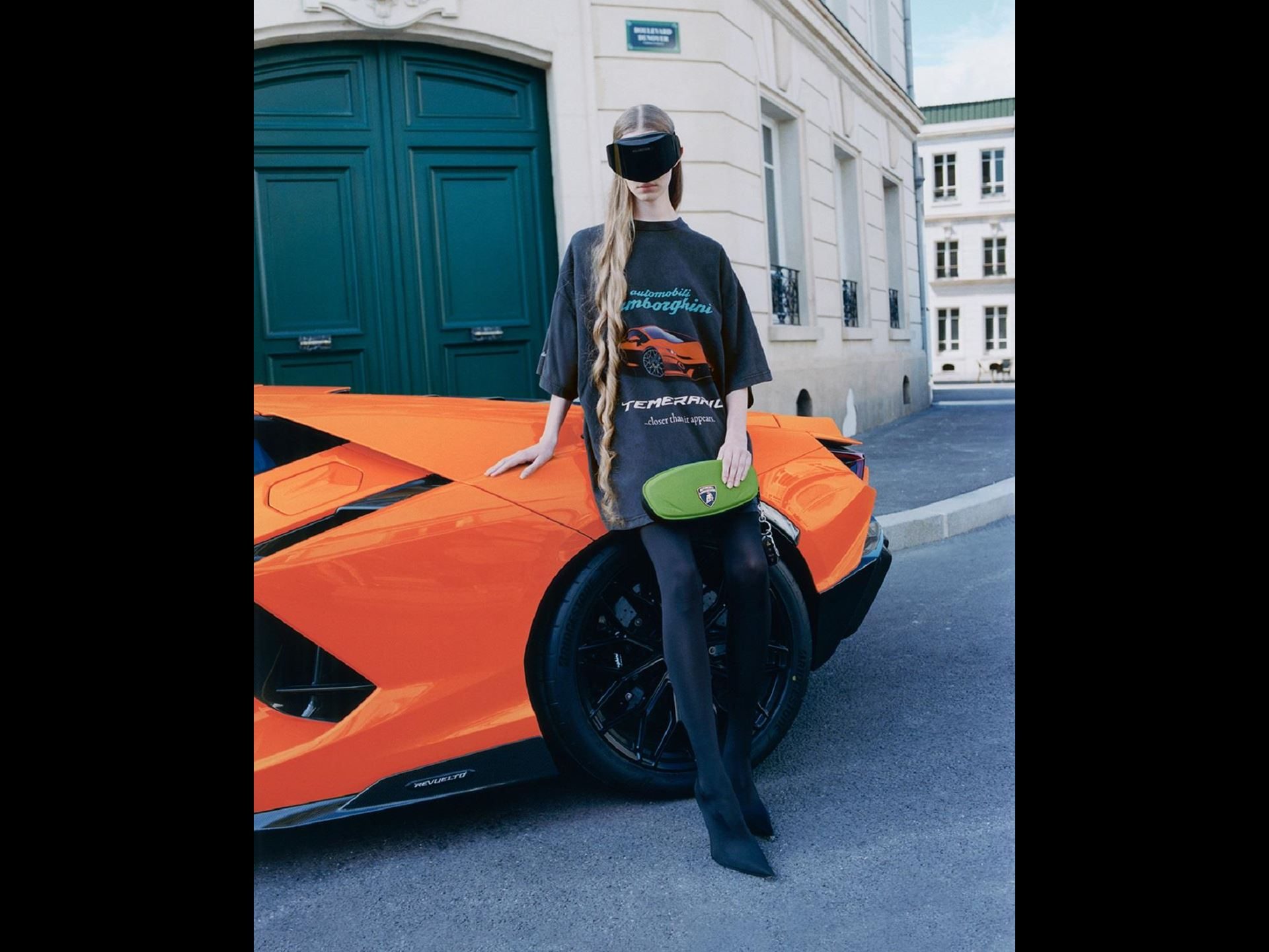 Balenciaga Lamborghini Collaboration Puts Temerario Artwork On The Runway And Graffiti-Wrapped Revueltos Outside Flagships Worldwide 5 Female model holding a green bag and wearing automobili lamborghini branded shirt, leaning on the rear of an orange lamborghini revuelto