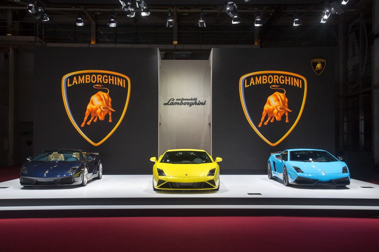 Three lamborghini gallardo models displayed on white platforms at the 2012 paris motorshow, including a dark blue convertible, a yellow coupe, and a light blue coupe