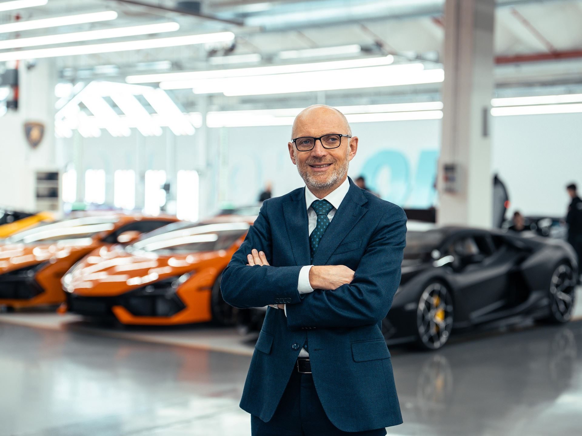 Lamborghini executive standing with arms crossed in front of a line of orange and black Revueltos on the factory floor
