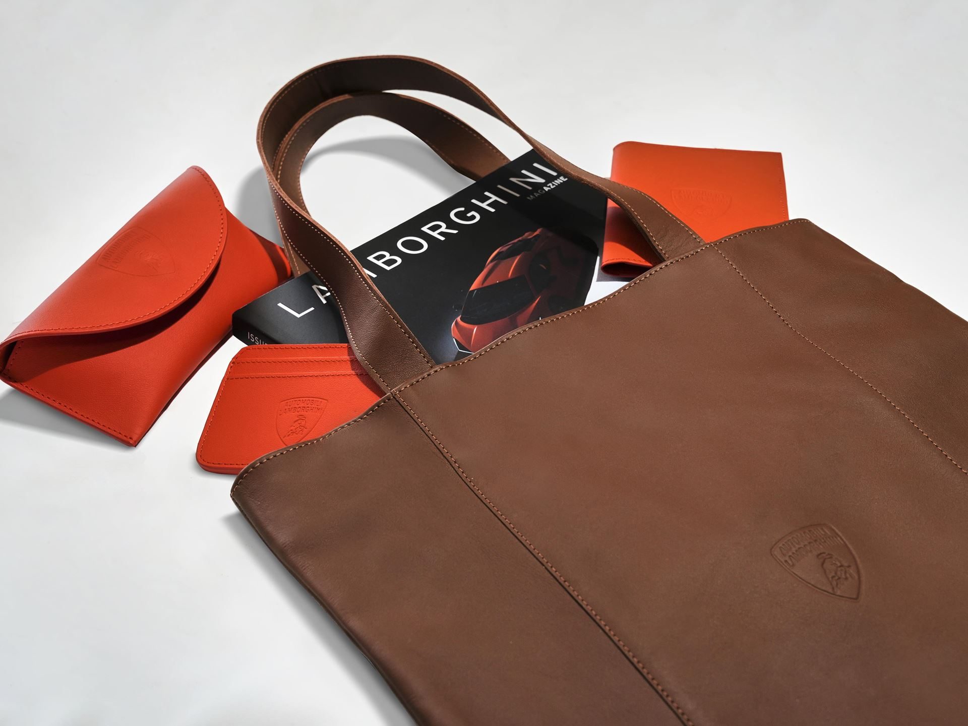 Lamborghini'S 2025 Holiday Gift Guide Stretches From Lego Bricks To $30,000 Loudspeakers 13 Brown leather lamborghini-branded tote bag with lamborghini magazine and smaller leather accessories on a white surface