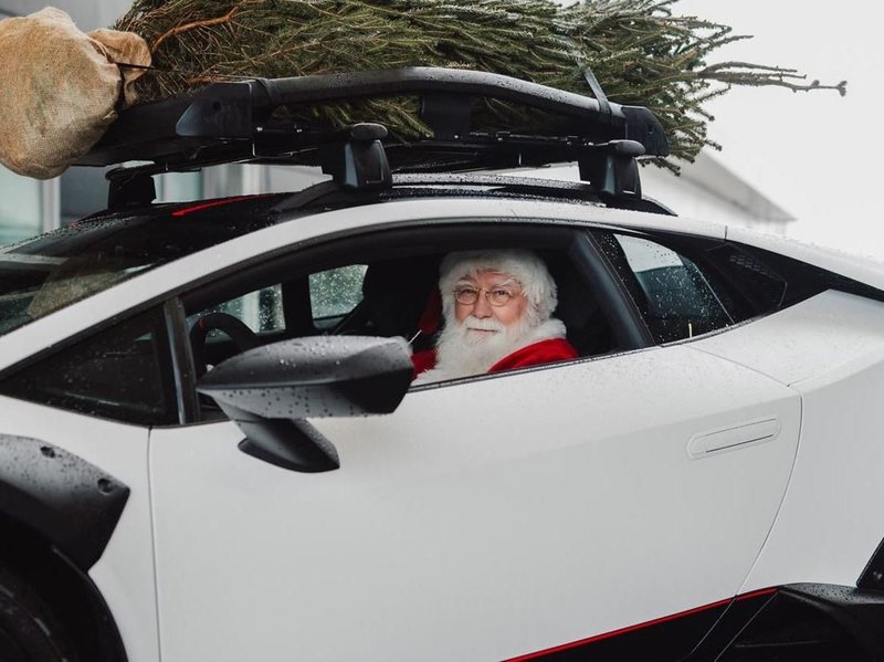 Santa Claus driving a white Lamborghini Huracán Sterrato with a Christmas tree strapped to the roof rack