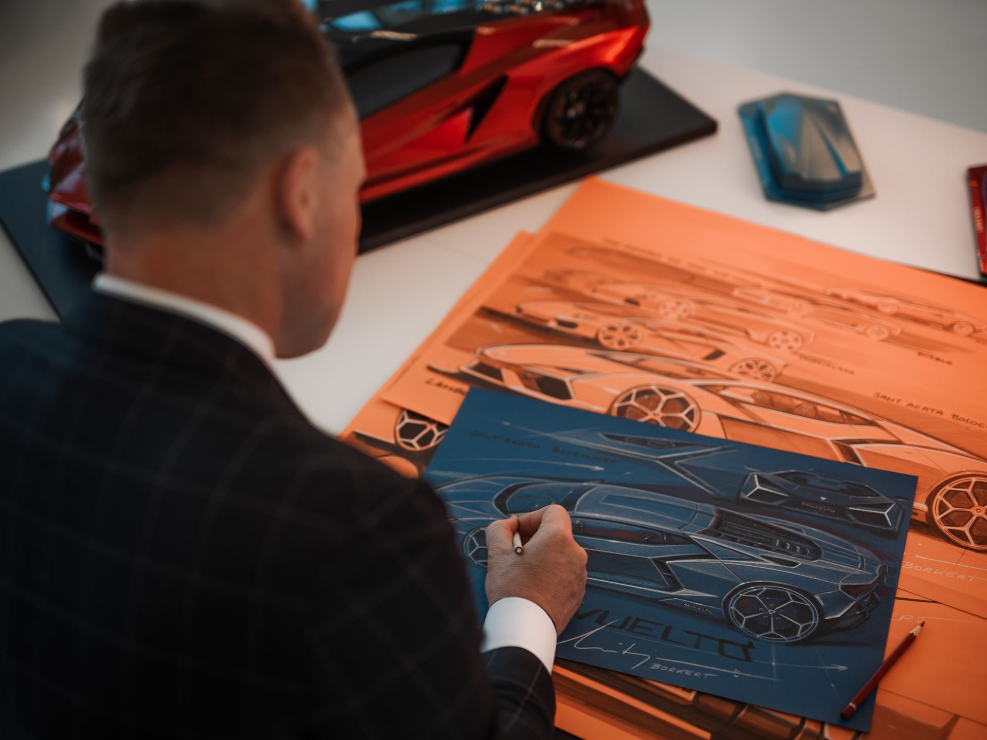 Every Lamborghini In The Lineup Just Won A Red Dot Award, And The Revuelto Took Top Honors 9 Overhead shot of a designer sketching a blue lamborghini revuelto concept on paper with a red model car nearby