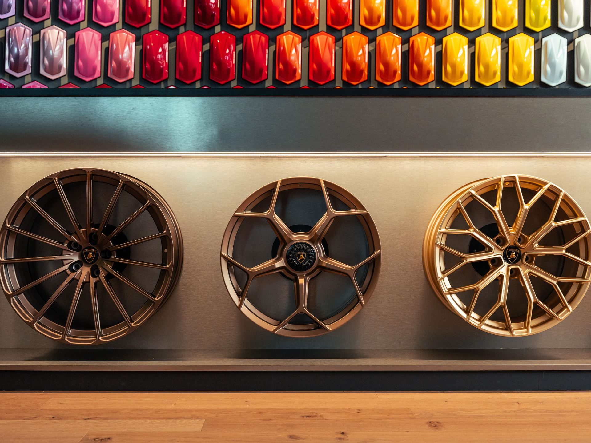 Why Lamborghini Names Its Paint Colors After Greek Gods And Diamond Dust 17 Three gold-colored lamborghini wheel designs displayed on a wall with paint samples visible above