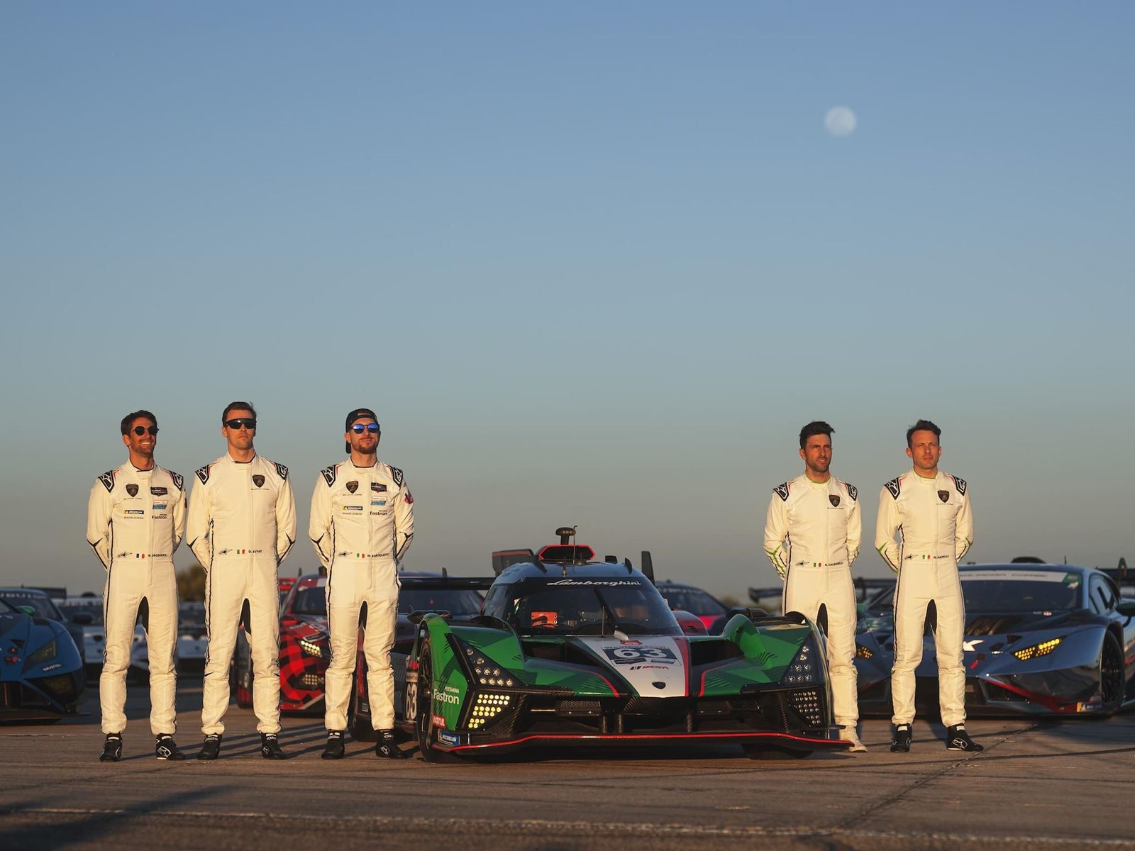 Six Lamborghini Squadra Corse drivers in white Alpinestars racing suits standing in front of the SC63 LMDh race car at the track