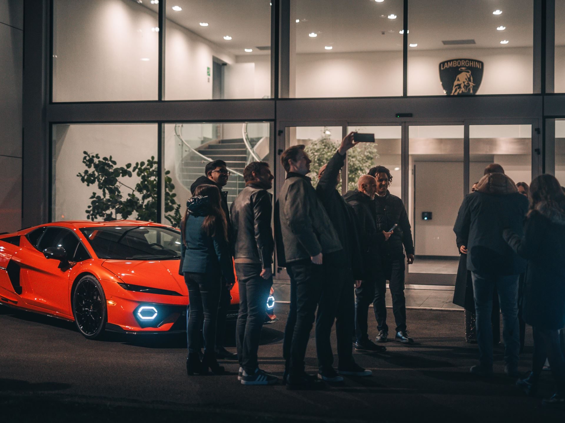 Lamborghini'S 2025 Christmas Video 'The Dinner' Puts Its Own Employees Center Stage 7 Orange lamborghini aventador parked outside a modern building with the lamborghini shield logo, surrounded by a group of people