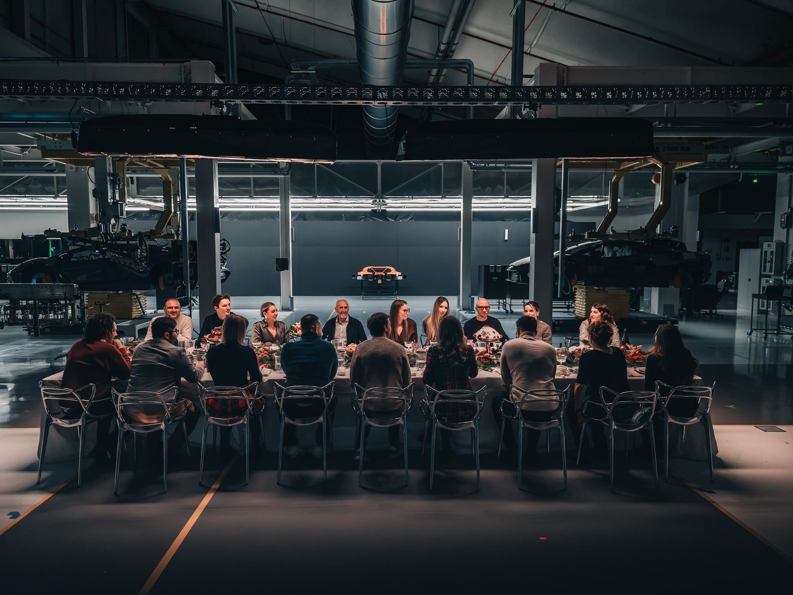 Lamborghini employees seated at a festively decorated dinner table inside a manufacturing facility, with an orange Lamborghini visible in the background