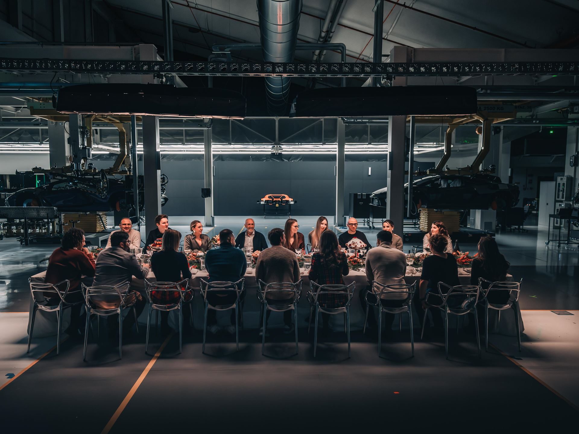 Lamborghini employees seated at a festively decorated dinner table inside a manufacturing facility, with an orange lamborghini visible in the background
