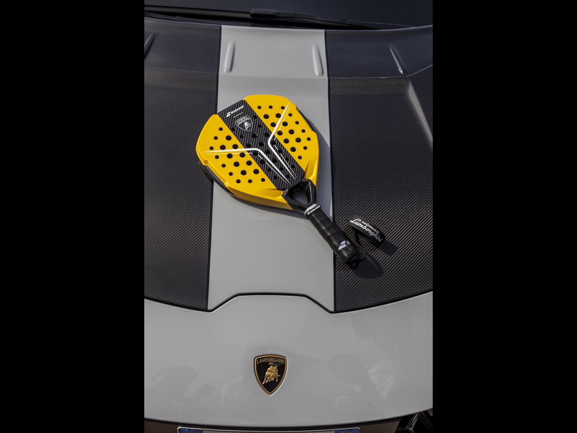 A yellow and black Babolat x Automobili Lamborghini padel racket resting on the hood of a grey Lamborghini car