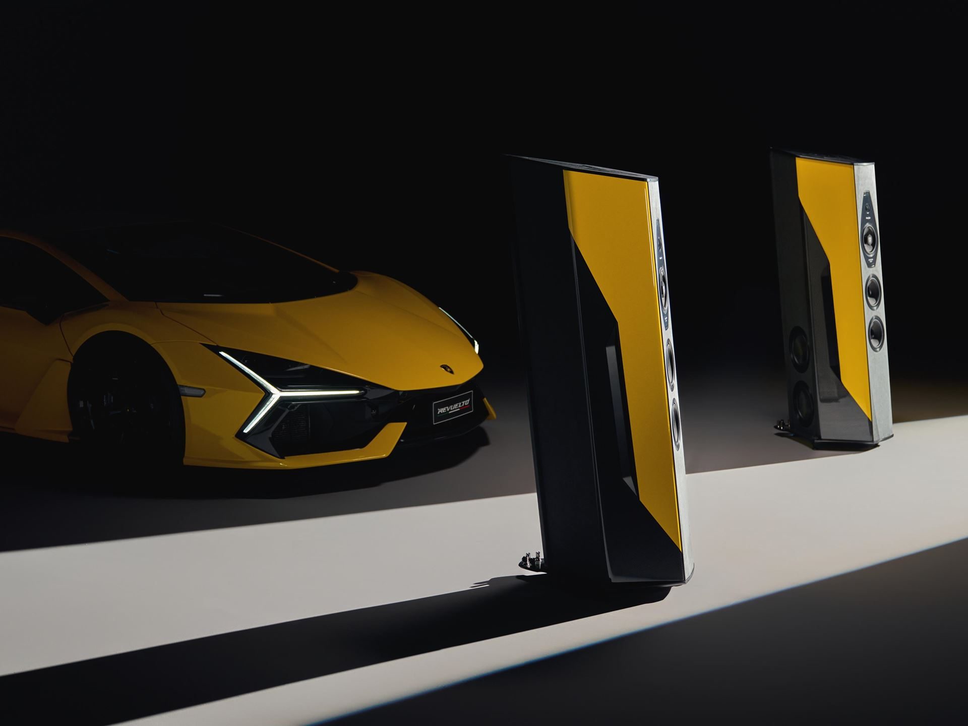 A yellow Lamborghini Revuelto partially visible behind two yellow and black Sonus Faber Il Cremonese Ex3me speakers
