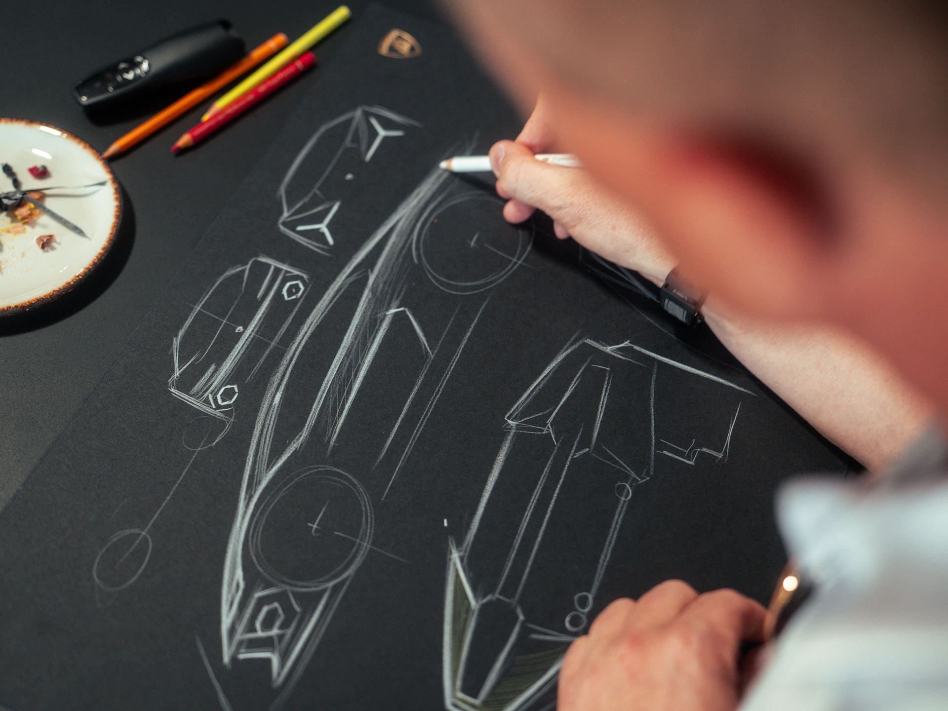 A designer's hands sketching angular Lamborghini concept forms and Y-shaped elements on black paper with white pencil