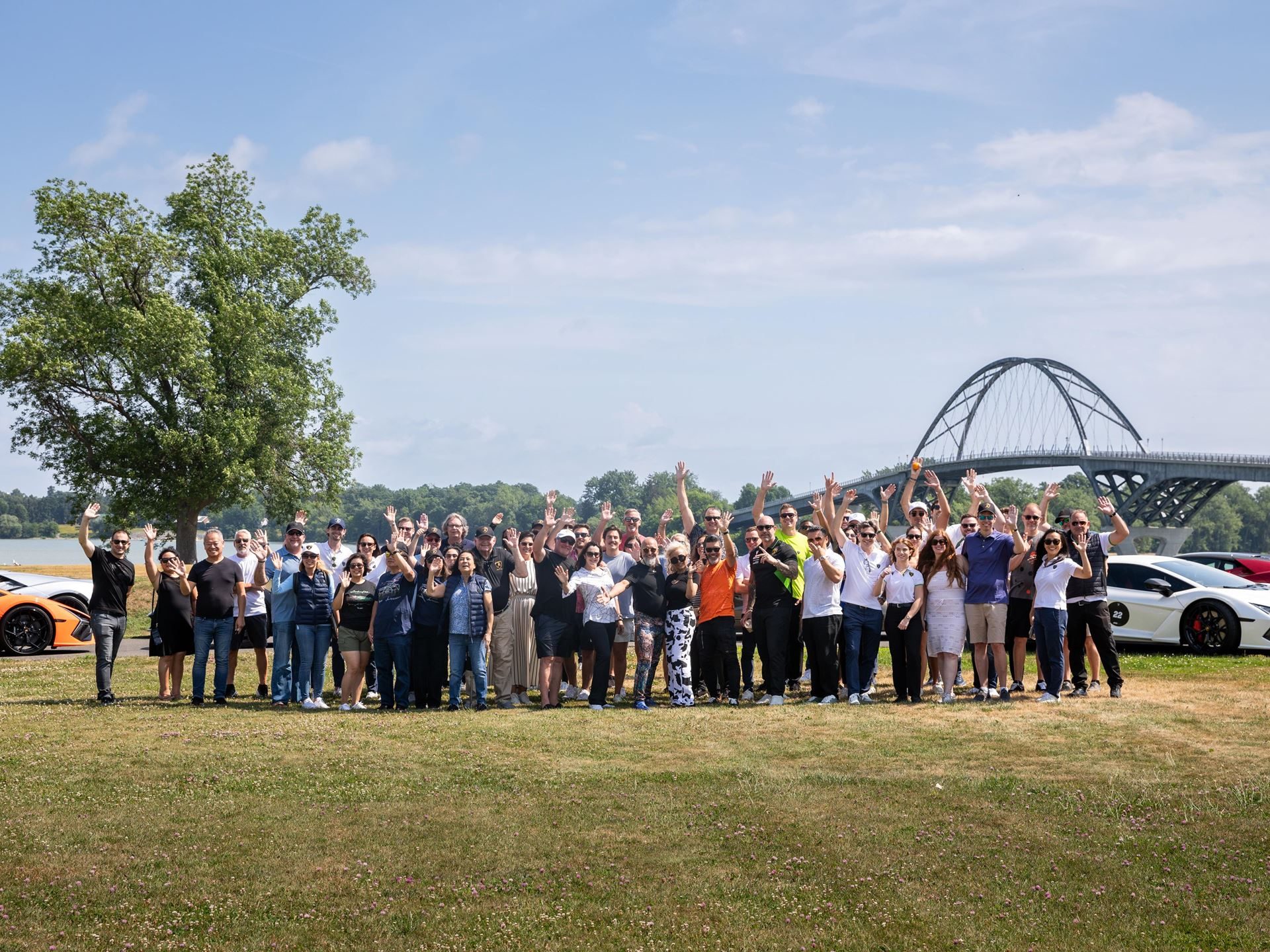 449 Miles Of New England Back Roads With 30 Lamborghinis: Inside The Esperienza Giro Usa 7 A large group of esperienza giro usa participants waves in a grassy field with lamborghinis and a bridge in the background