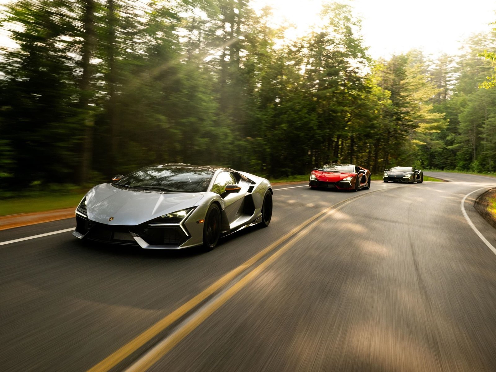 A silver Lamborghini Revuelto leads a convoy of supercars on a winding New England forest road