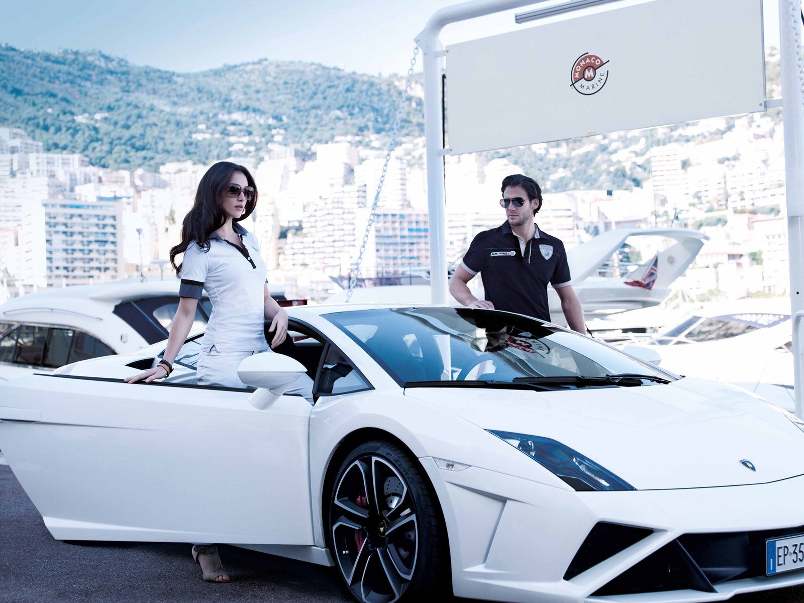 White Lamborghini Gallardo parked at a marina with two people posing beside it, yachts and mountains in the background
