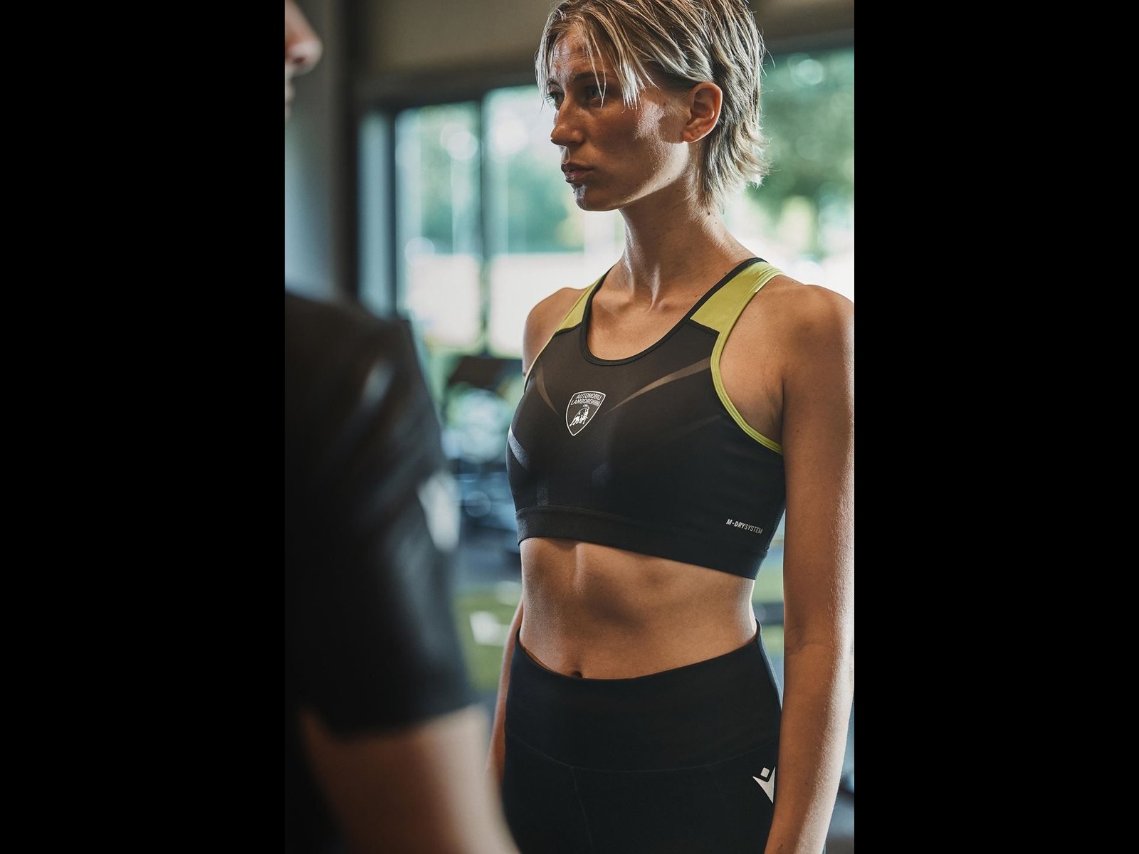 Close-up of female model wearing black Lamborghini-branded Macron sports bra with neon yellow accents and shield logo