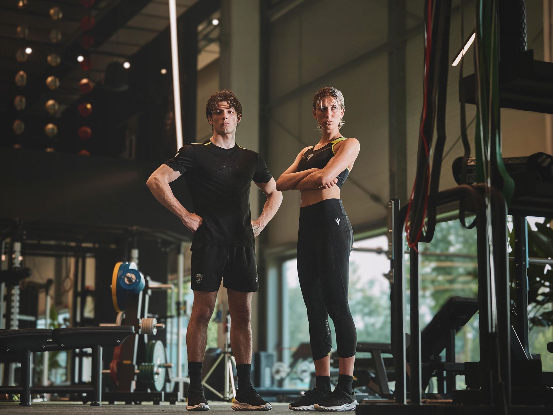 Lamborghini And Macron'S Second Activewear Drop Brings Sant'Agata'S Dna Into The Gym 9 Male and female models standing in gym wearing black lamborghini-branded macron activewear with yellow accents and shield logo