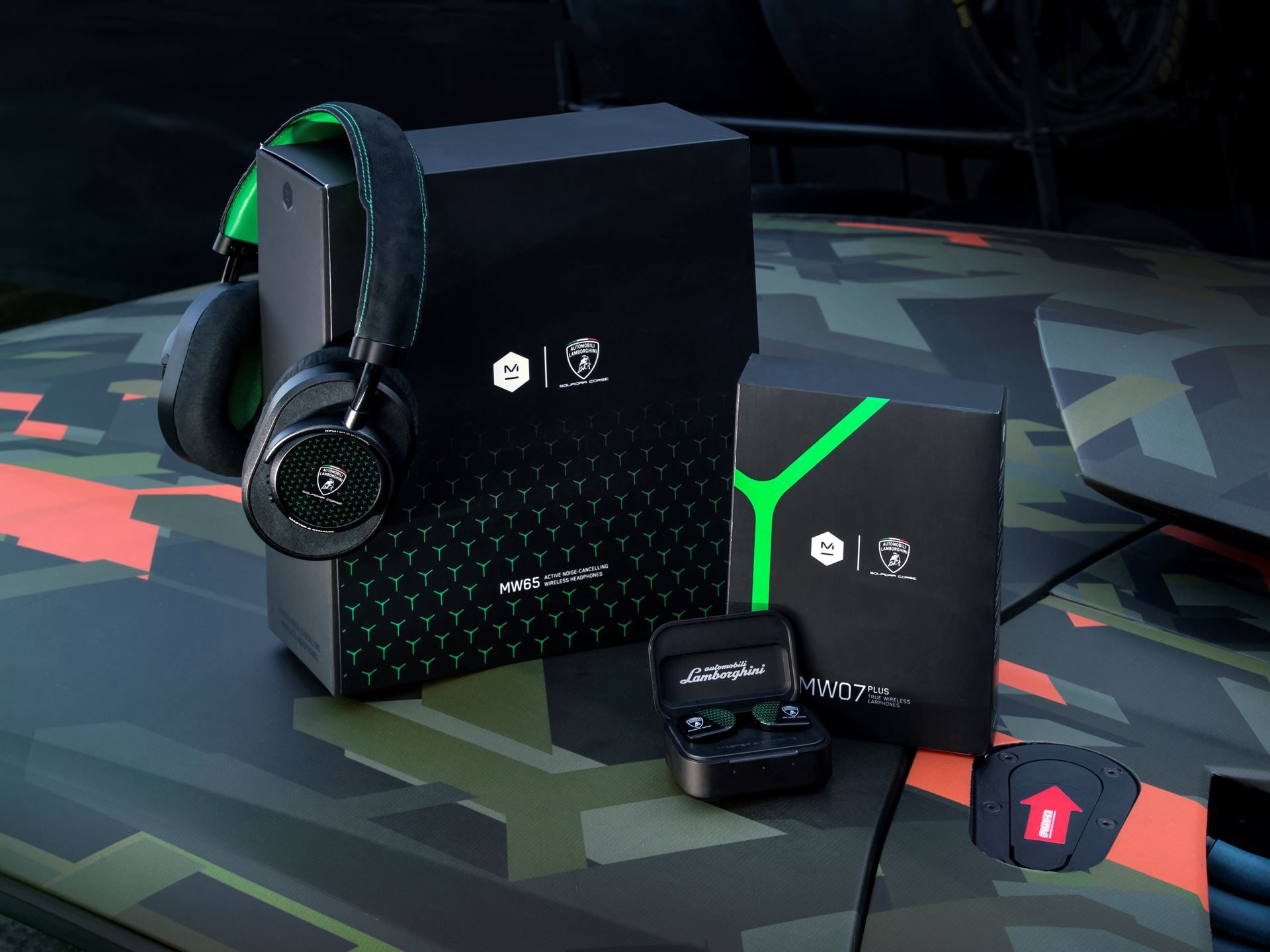 Master & dynamic x automobili lamborghini headphones and earbuds with packaging displayed on the camouflage-patterned hood of a lamborghini vehicle, showing lime green accents