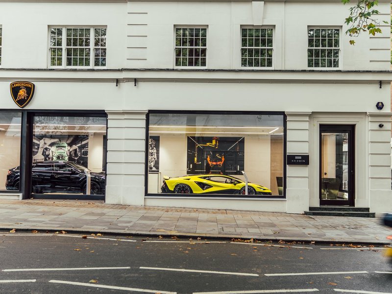 Exterior view of Lamborghini Mayfair showroom in Berkeley Square, with a black Urus SE and yellow Revuelto visible through large glass windows