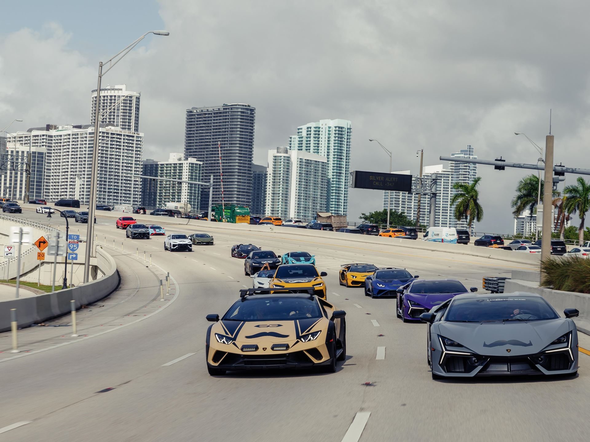 150 Mustache-Clad Lamborghinis Hit Miami As Movember Campaign Enters Year Four 5 A convoy of lamborghinis including a gold huracán sterrato and grey revuelto driving on a highway with mustache decals and a city skyline behind them