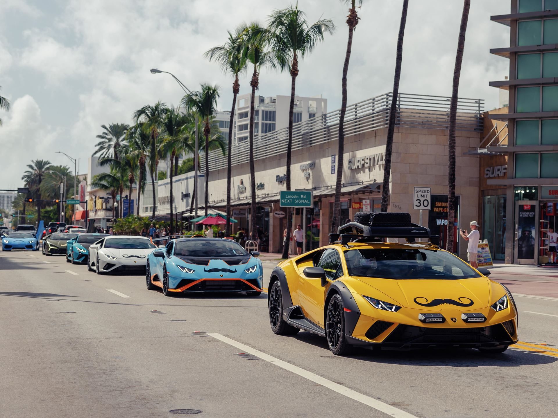 150 Mustache-Clad Lamborghinis Hit Miami As Movember Campaign Enters Year Four 11 A line of lamborghini huracáns including a yellow sterrato with roof rack driving down a palm-tree lined street with mustache decals