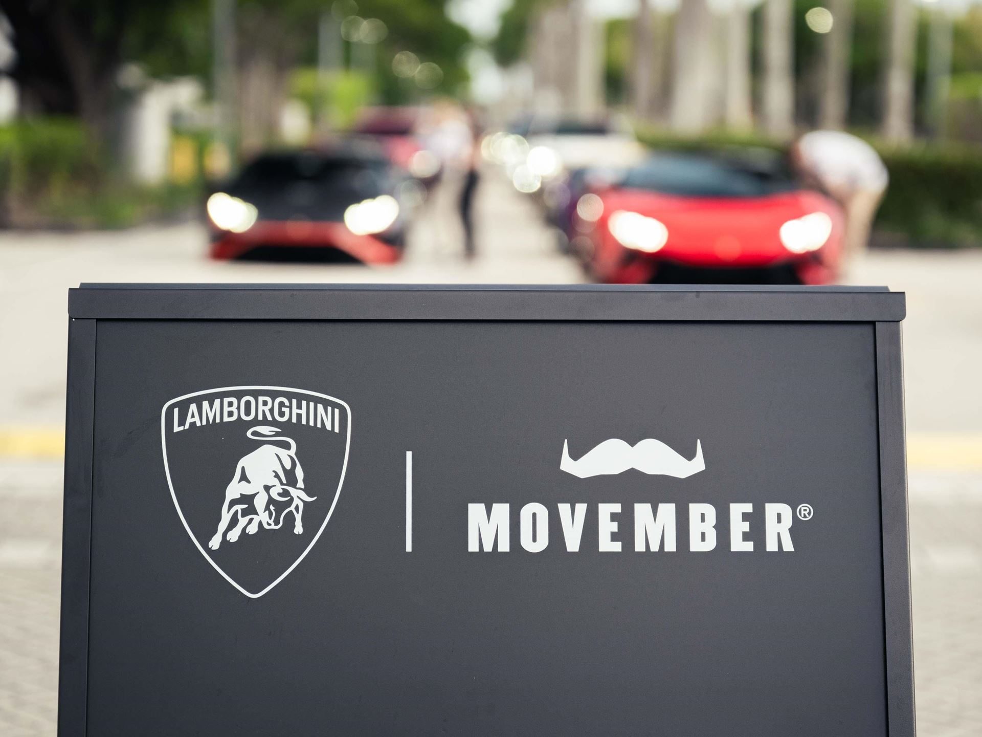 150 Mustache-Clad Lamborghinis Hit Miami As Movember Campaign Enters Year Four 3 A black sign displaying the lamborghini logo alongside the movember logo with blurred lamborghini cars in the background
