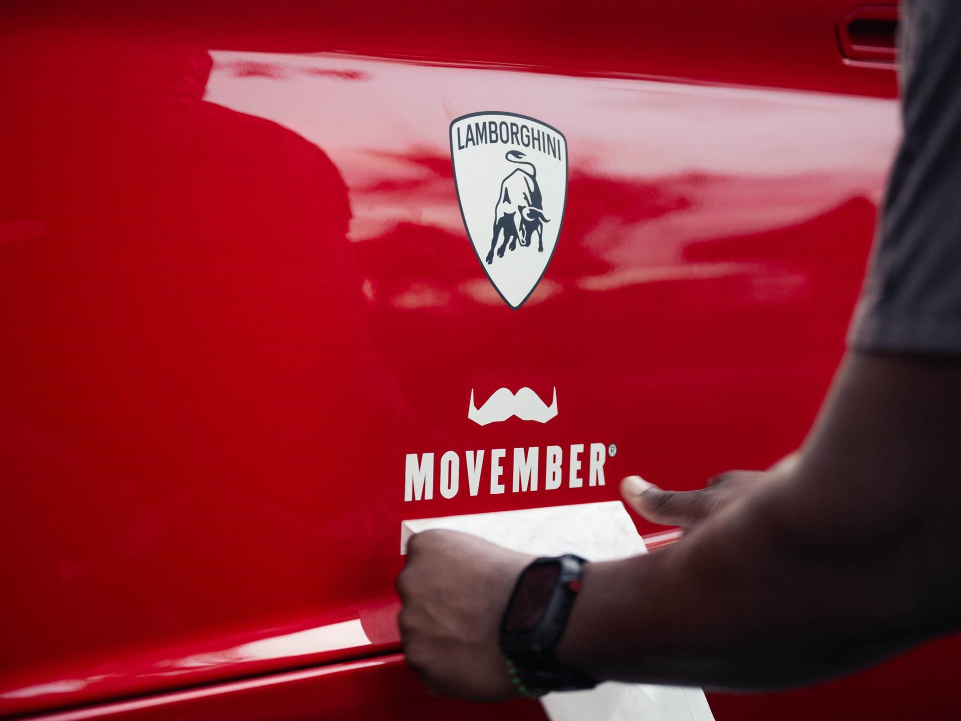 150 Mustache-Clad Lamborghinis Hit Miami As Movember Campaign Enters Year Four 7 A person applying a movember decal with a mustache logo and lamborghini shield to the side of a red lamborghini