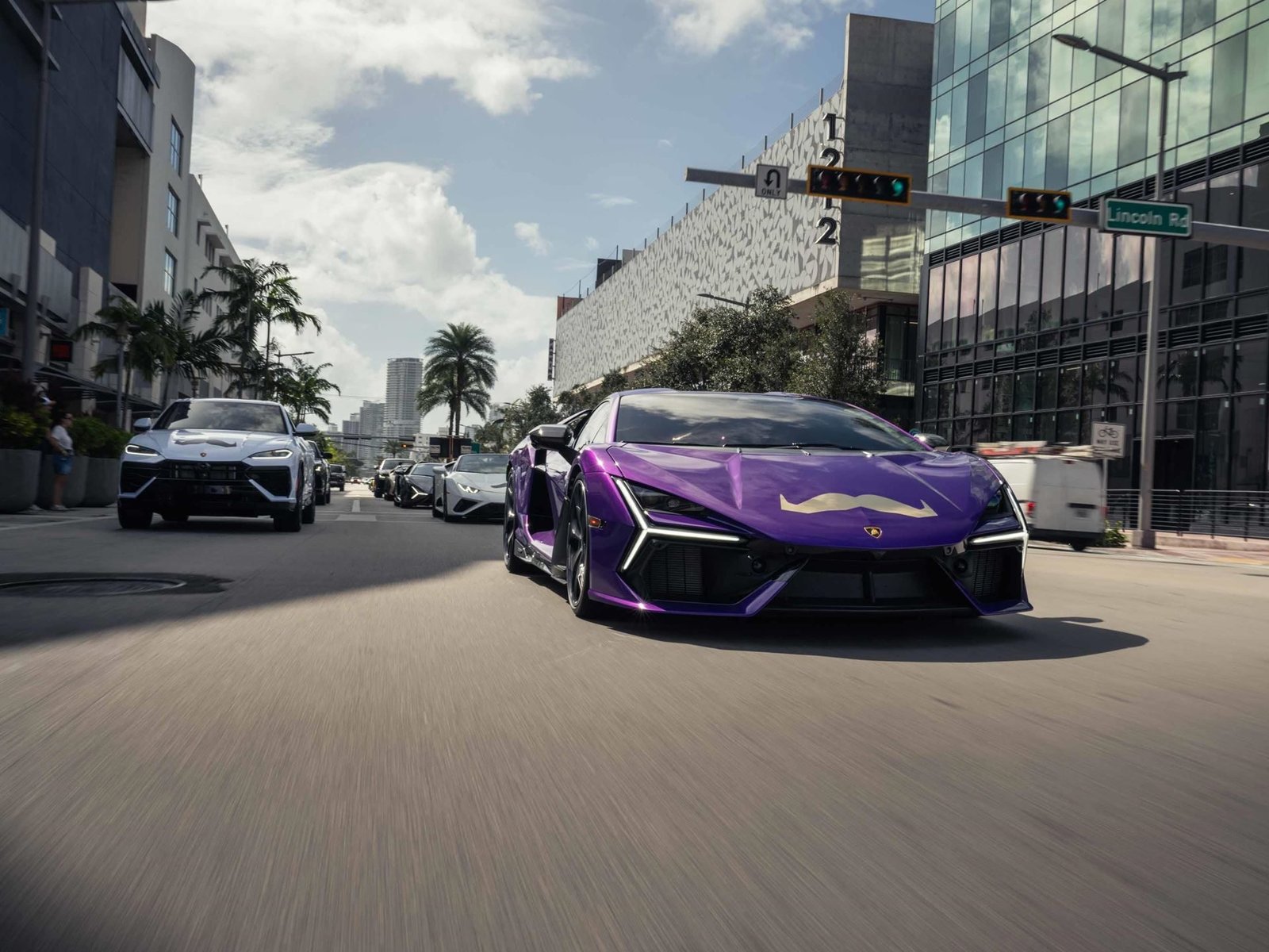 A purple Lamborghini Sián FKP 37 with a Movember mustache decal leads a convoy of Lamborghinis through a city street