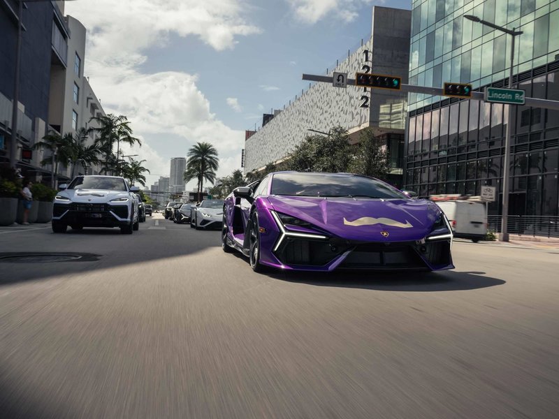 A purple Lamborghini Sián FKP 37 with a Movember mustache decal leads a convoy of Lamborghinis through a city street